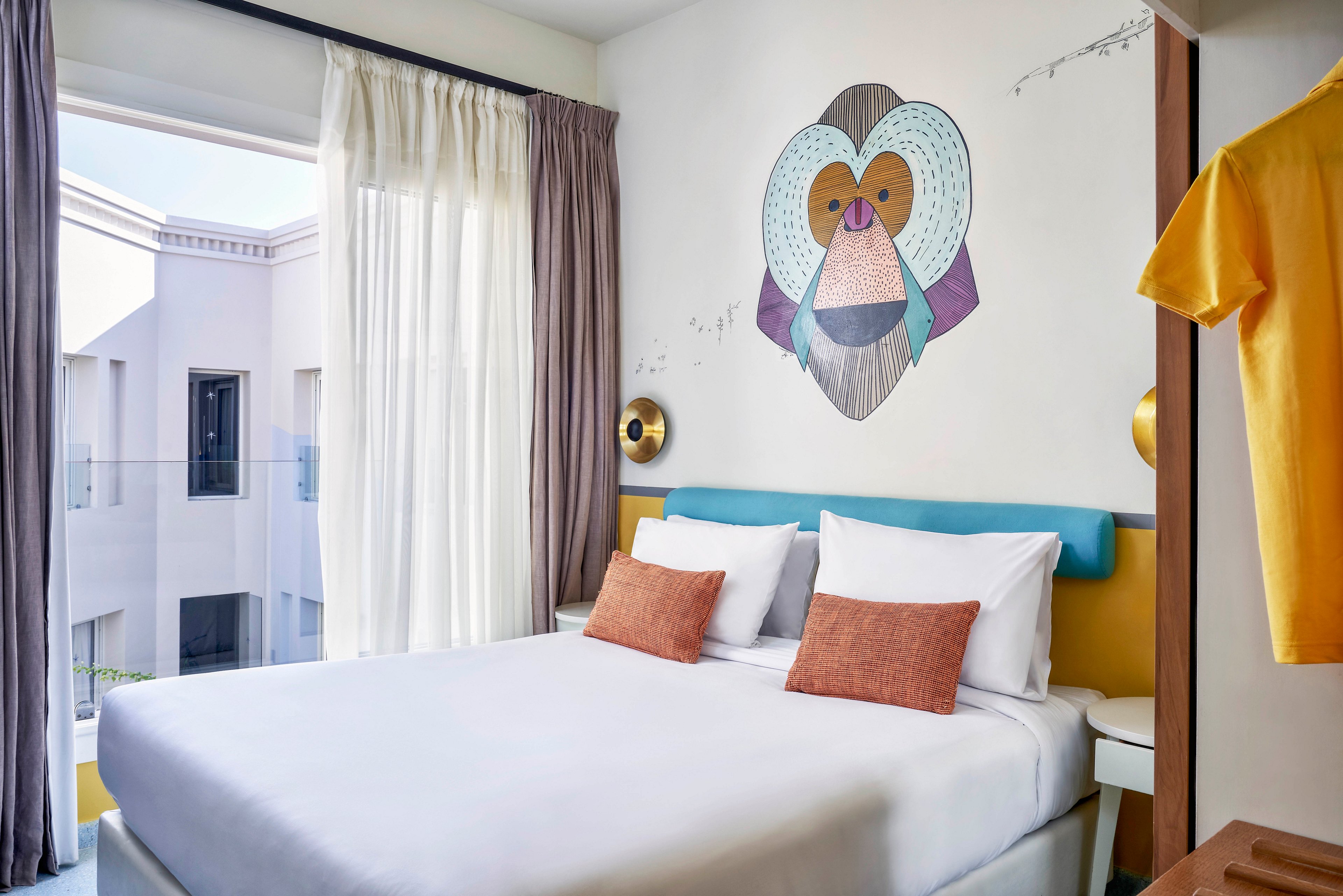 A contemporary hotel room features a large bed with white linens and orange accent pillows. The wall above the bed displays a colorful, stylized monkey face mural. The room is bright with natural light from a balcony, and soft curtains frame the window. A yellow shirt hangs in the foreground, adding a pop of color to the neutral palette.