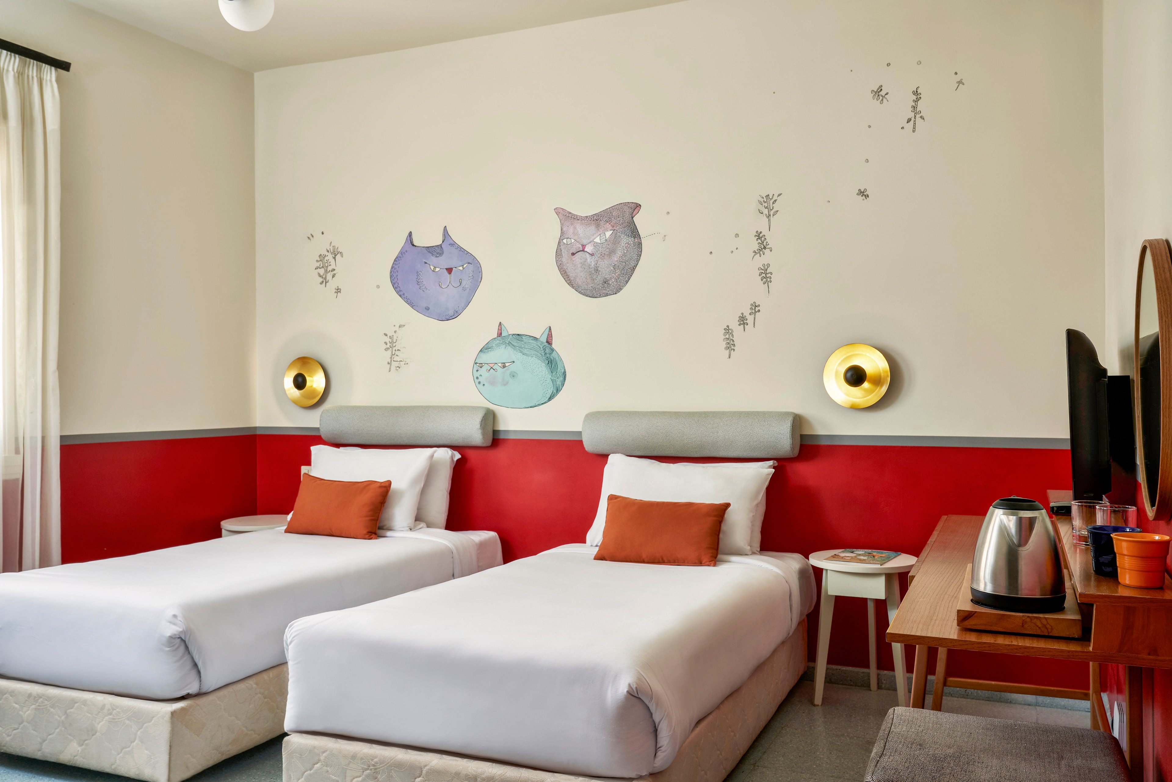 A contemporary hotel room features two single beds with white linens and orange accent pillows. The wall above the beds displays playful cat illustrations in pastel colors, adding a whimsical touch. The room includes a wooden desk, electric kettle, and round mirrors, with soft lighting and a red and white color palette.