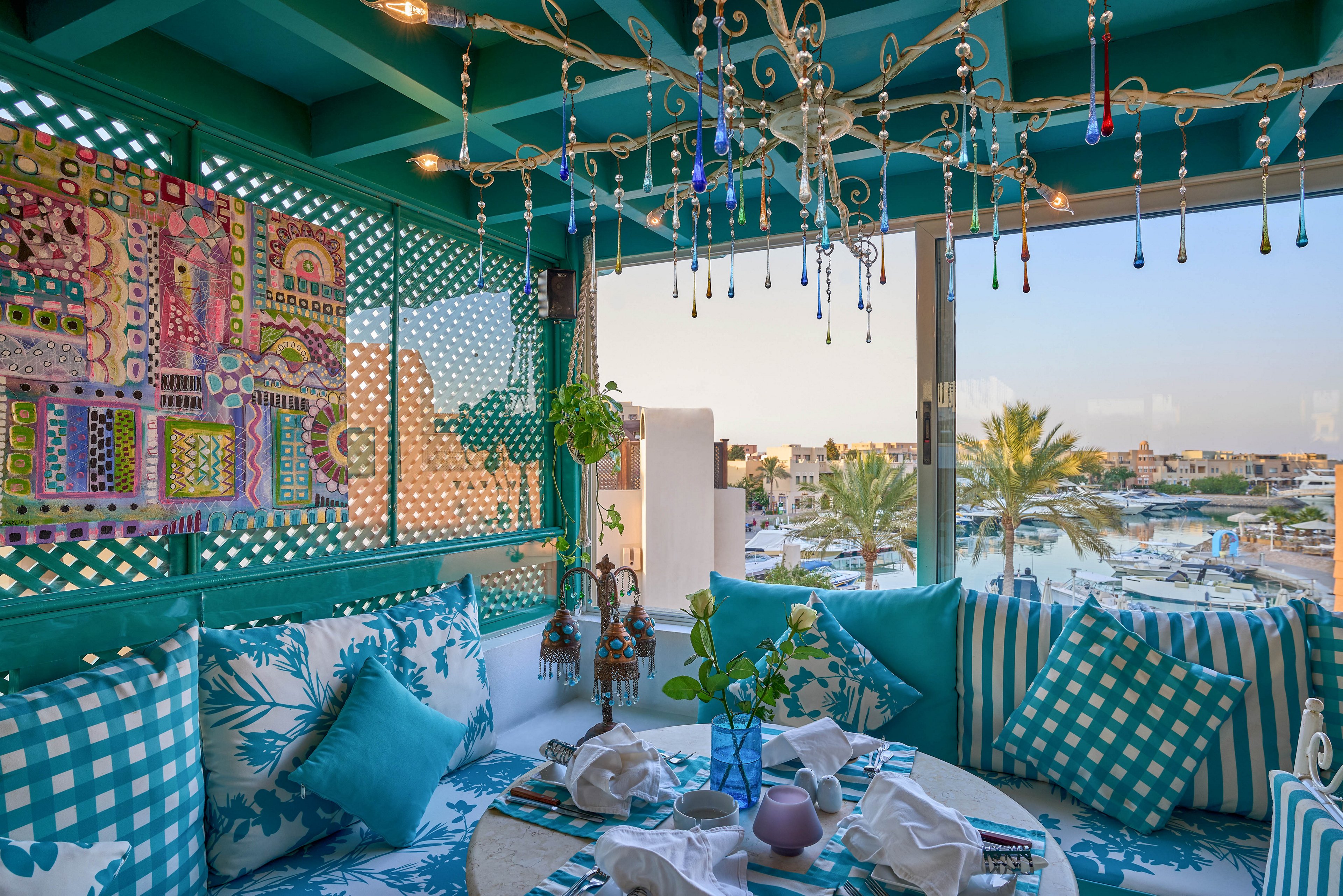 A vibrant terrace lounge features turquoise and white patterned cushions, a round table set for dining, and eclectic wall art. The setting overlooks a marina with palm trees and boats, visible through large windows. The mood is relaxed and inviting, with a Mediterranean coastal style and a bright, airy palette.