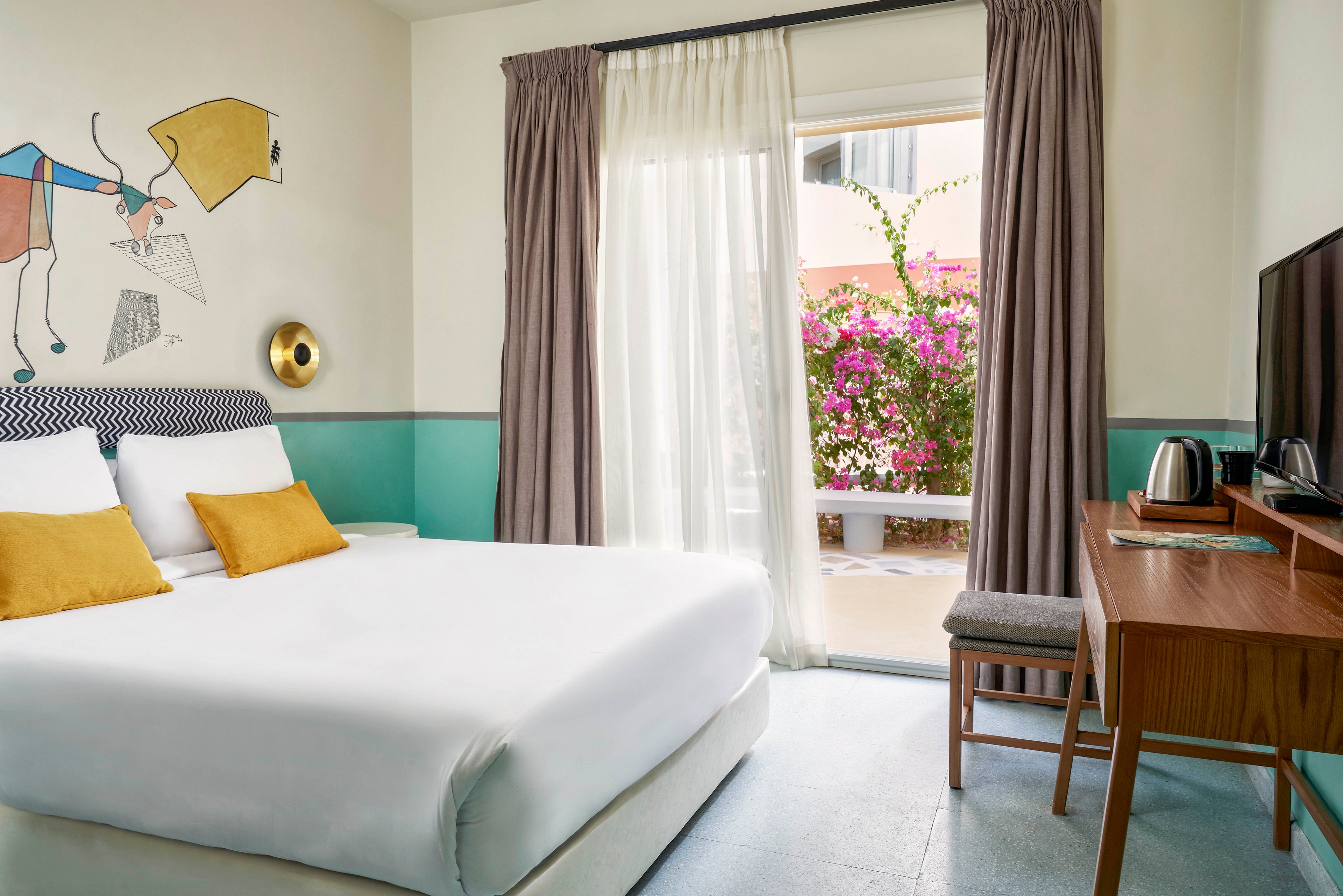 A bright, contemporary hotel room features a large bed with white linens and yellow accent pillows. The space includes a desk with a kettle and cups, a television, and a window with sheer curtains overlooking a blooming garden with pink flowers. The walls are decorated with playful abstract art and soft pastel colors, creating a calm and inviting atmosphere.