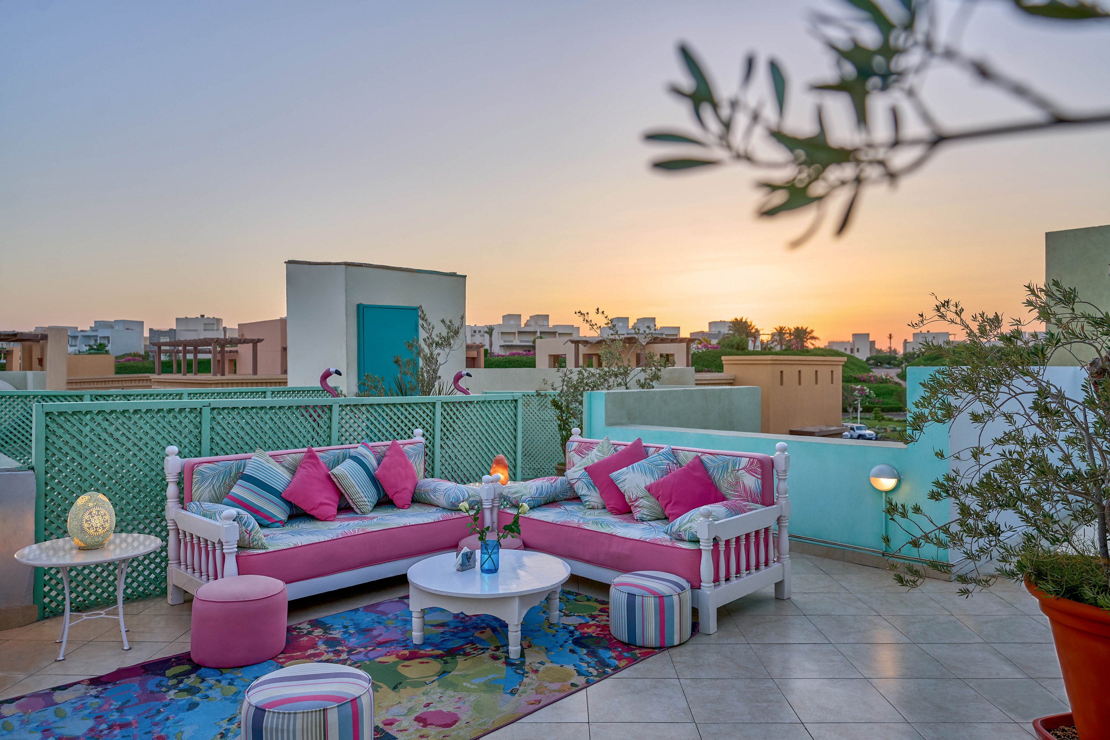A vibrant rooftop terrace features two white wooden sofas with pink and blue cushions, arranged around a small white coffee table. The setting is enhanced by patterned rugs, striped ottomans, and decorative plants, creating a cozy and inviting atmosphere. The sun sets in the background, casting a warm glow over the urban skyline. The overall mood is relaxed and cheerful, with a playful mix of colors and patterns.
