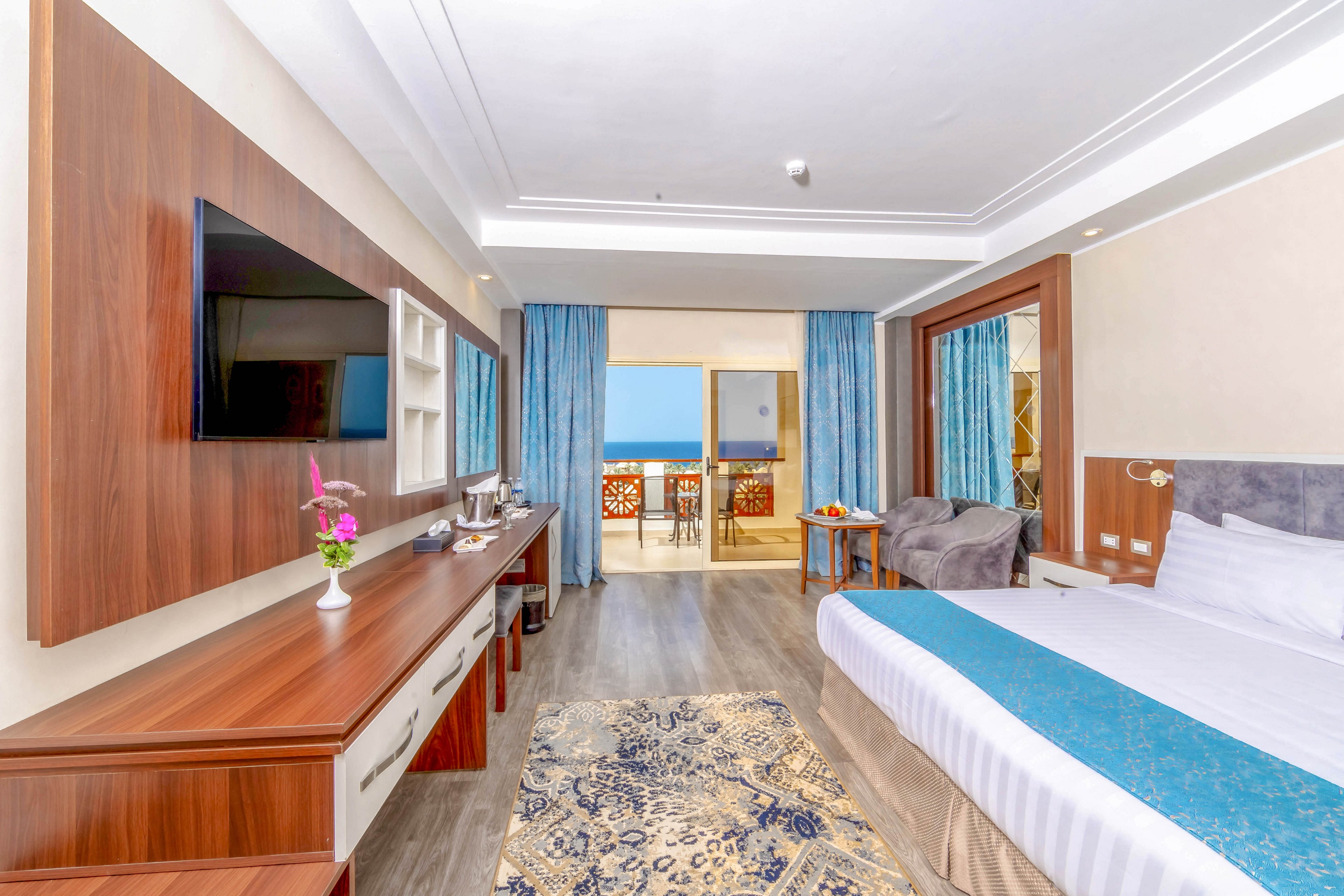 A spacious and well-lit hotel room featuring a large bed with blue accents, a flat-screen TV, and a wooden desk. The room opens to a balcony with a view of the ocean, complemented by blue curtains and a cozy seating area. A decorative rug and fresh flowers add a touch of elegance to the space.