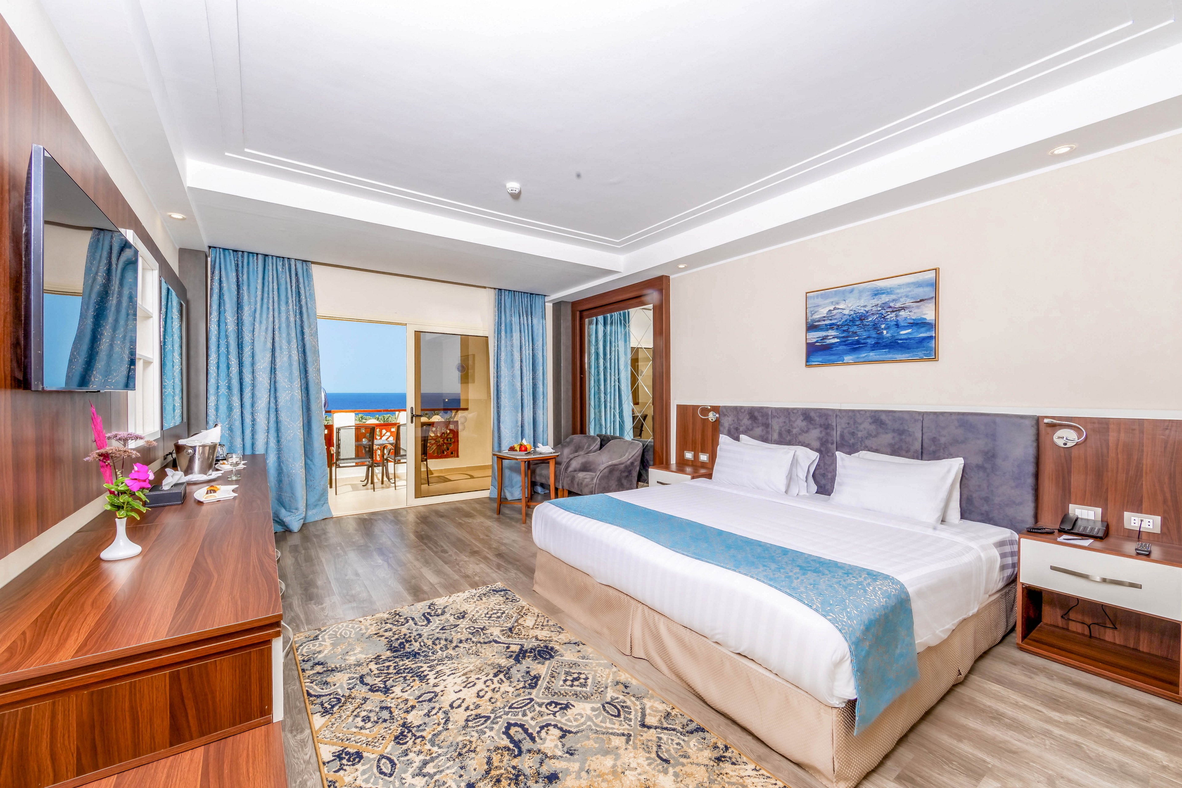 A spacious and modern hotel room featuring a large bed with white and blue bedding. The room includes a wooden desk, a flat-screen TV, and a seating area with a view of the ocean through sliding glass doors. Decorative elements include a patterned rug, wall art, and fresh flowers on the desk. The room is well-lit with natural light and has a serene ambiance.