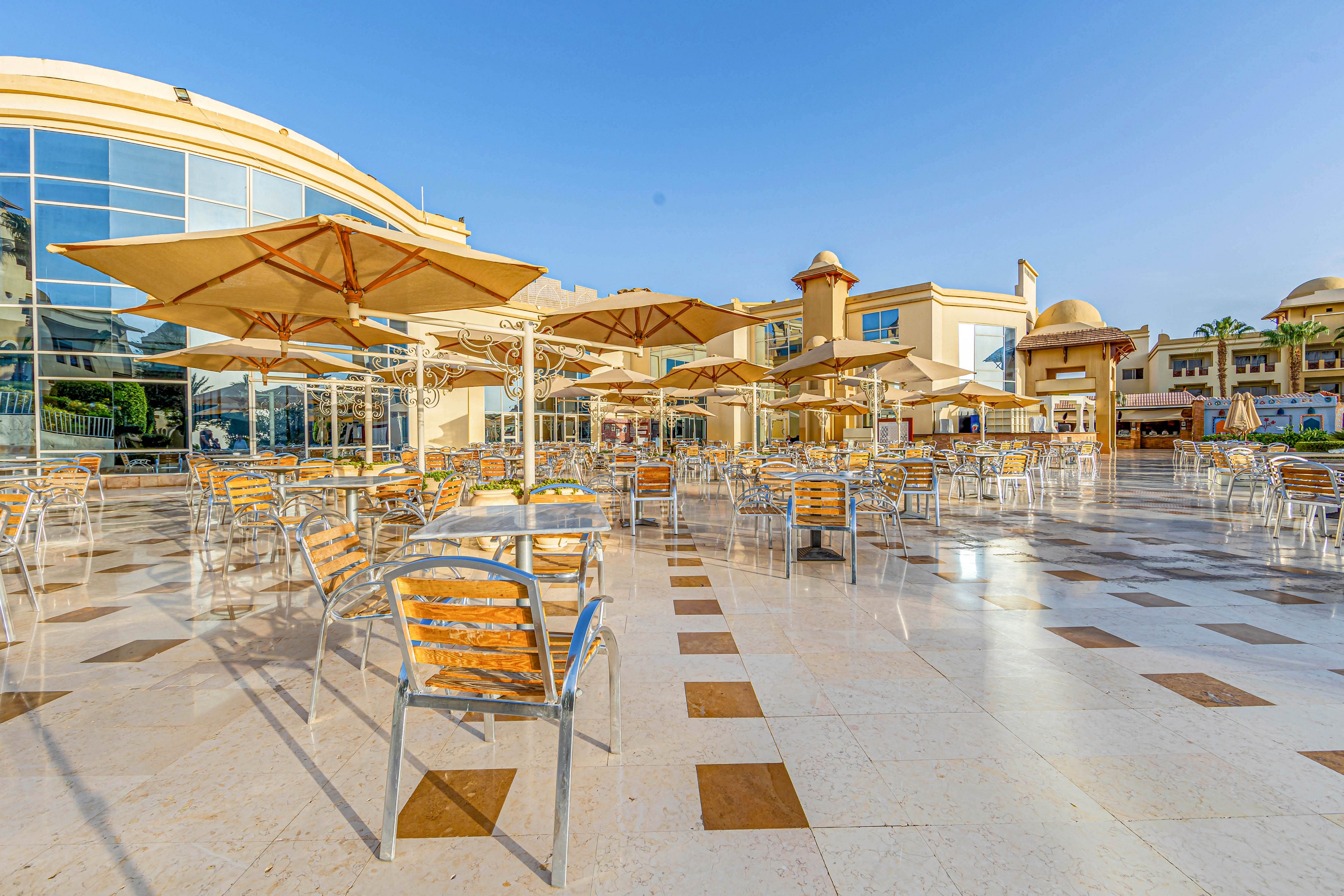 An outdoor cafe area featuring numerous wooden and metal chairs and tables under large beige umbrellas. The setting is a sunny day with a clear blue sky, and the cafe is surrounded by modern architecture with glass windows and domed structures. The tiled flooring adds a clean and polished look to the space.
