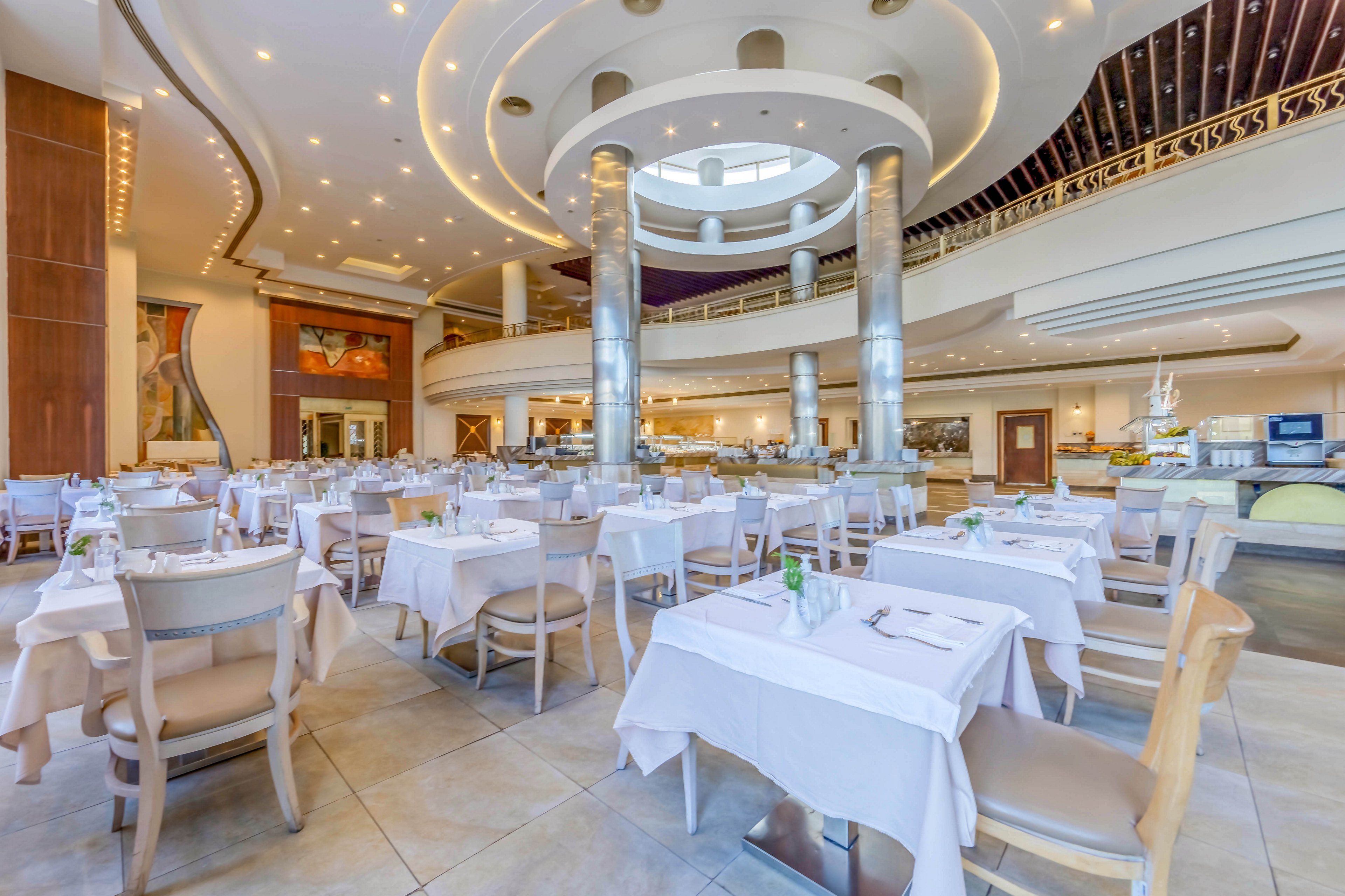 A spacious and well-lit restaurant interior featuring white tablecloths, neatly arranged tables, and modern architectural elements. The setting includes a high ceiling with circular designs, polished columns, and warm ambient lighting. The atmosphere is clean and inviting, ideal for dining or special events.