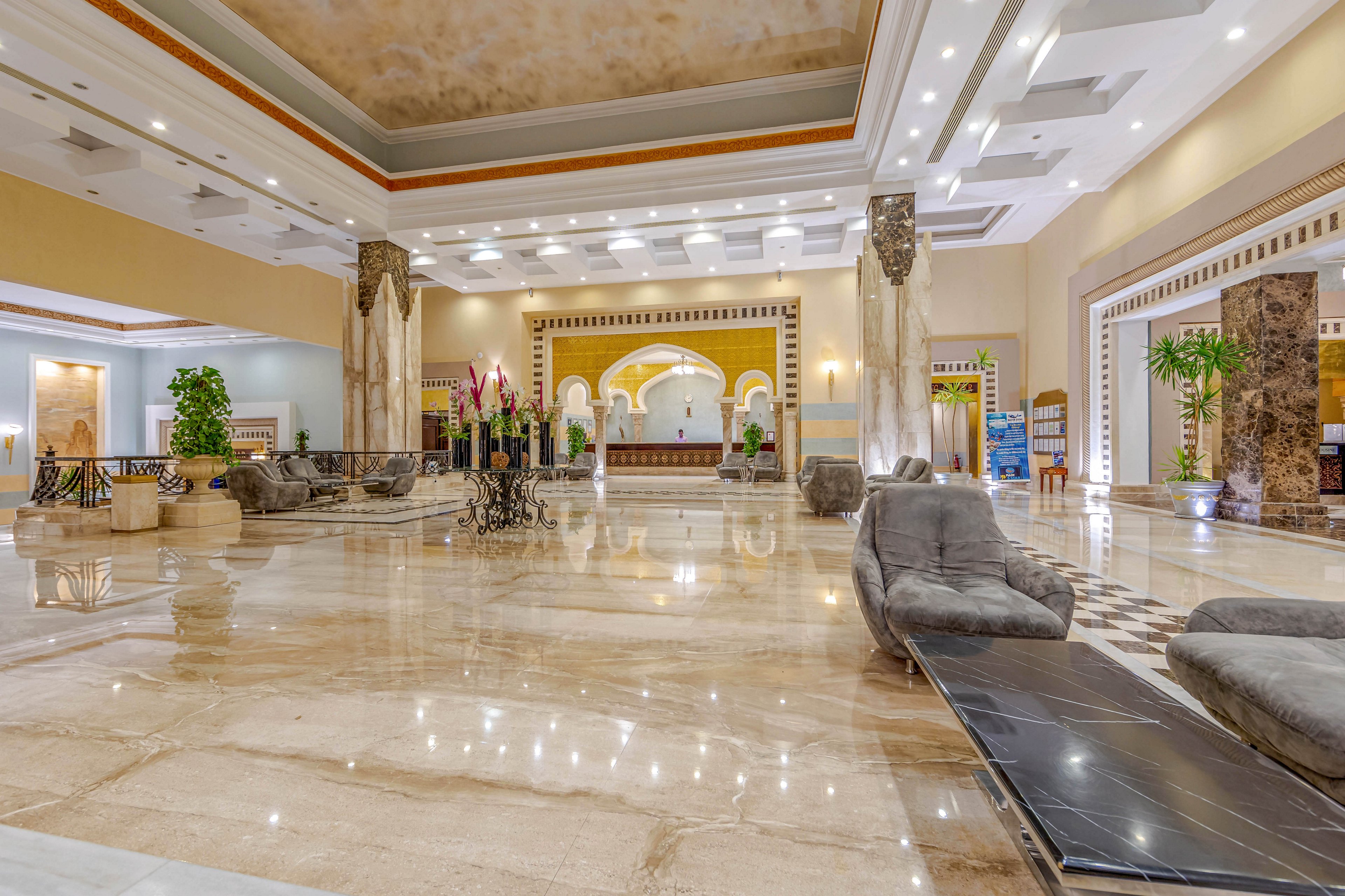A grand and spacious hotel lobby featuring polished marble floors, elegant seating arrangements, and decorative columns. The setting is well-lit with warm lighting and includes greenery and ornate architectural details. The ambiance exudes sophistication and comfort, ideal for relaxation or social gatherings.