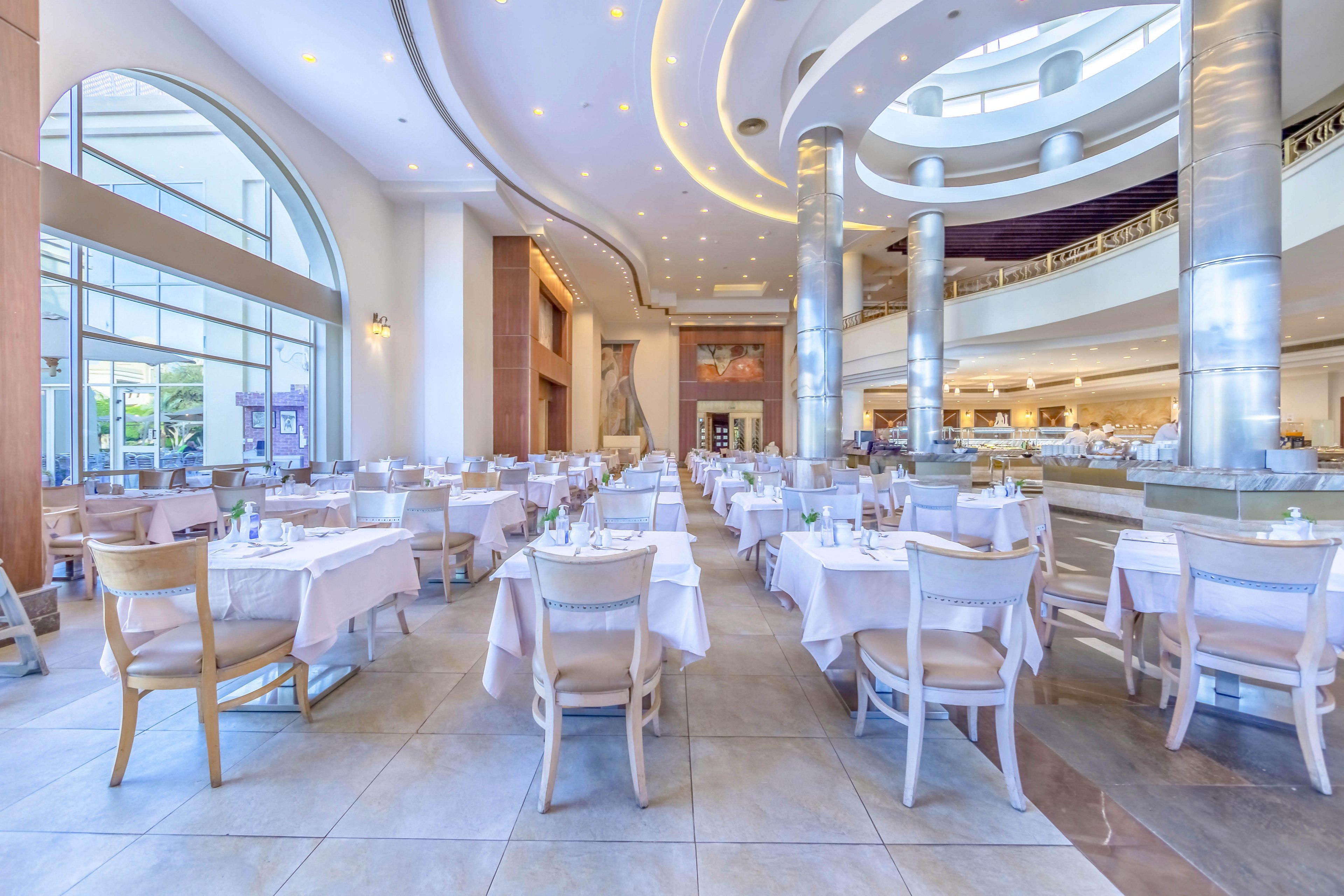 A spacious and well-lit restaurant interior featuring neatly arranged tables with white tablecloths and wooden chairs. The setting includes high ceilings, large arched windows, and contemporary architectural details with metallic accents. The atmosphere is bright and inviting, ideal for dining or events.