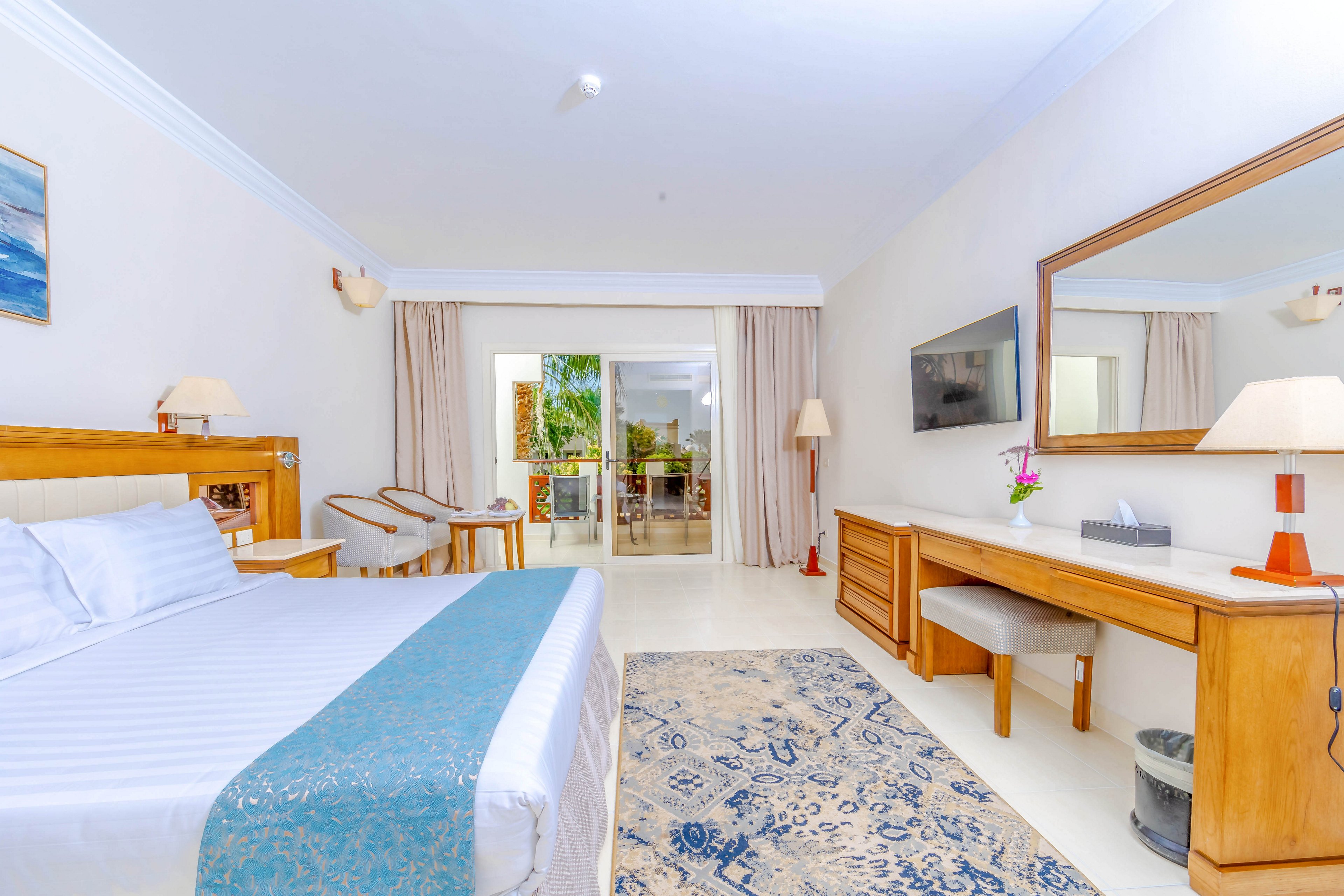 A spacious and well-lit hotel room featuring a large bed with a blue accent runner, wooden furniture, and a wall-mounted TV. The room includes a desk, chairs, and a mirror, with a sliding glass door leading to a balcony overlooking greenery. The neutral color palette and natural light create a relaxing and inviting atmosphere.