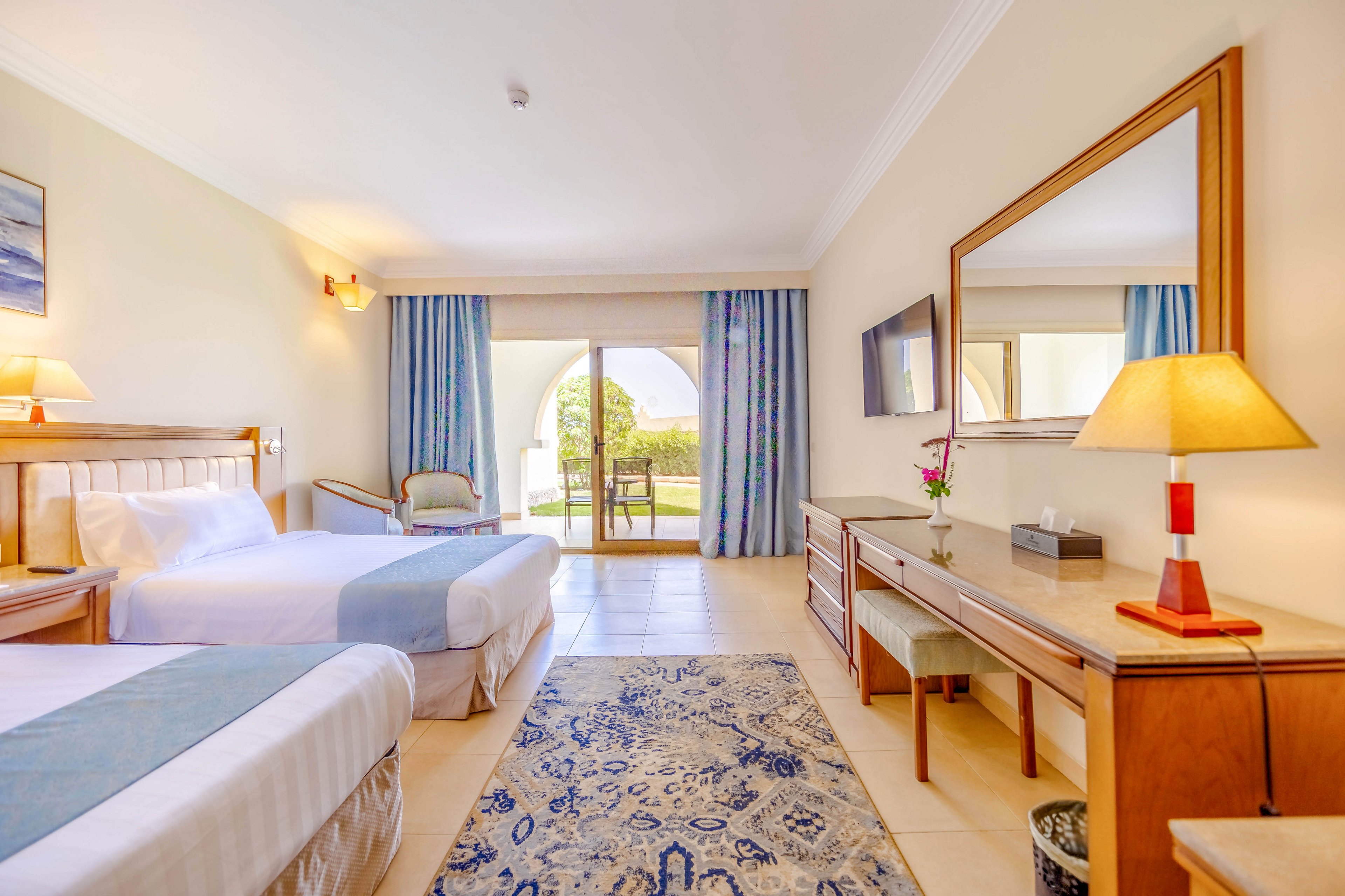 A spacious and well-lit hotel room featuring two neatly made twin beds with light blue accents. The room includes a wooden desk, a large mirror, and a flat-screen TV mounted on the wall. A sliding glass door opens to a patio with a garden view, complemented by soft natural light and neutral decor tones.
