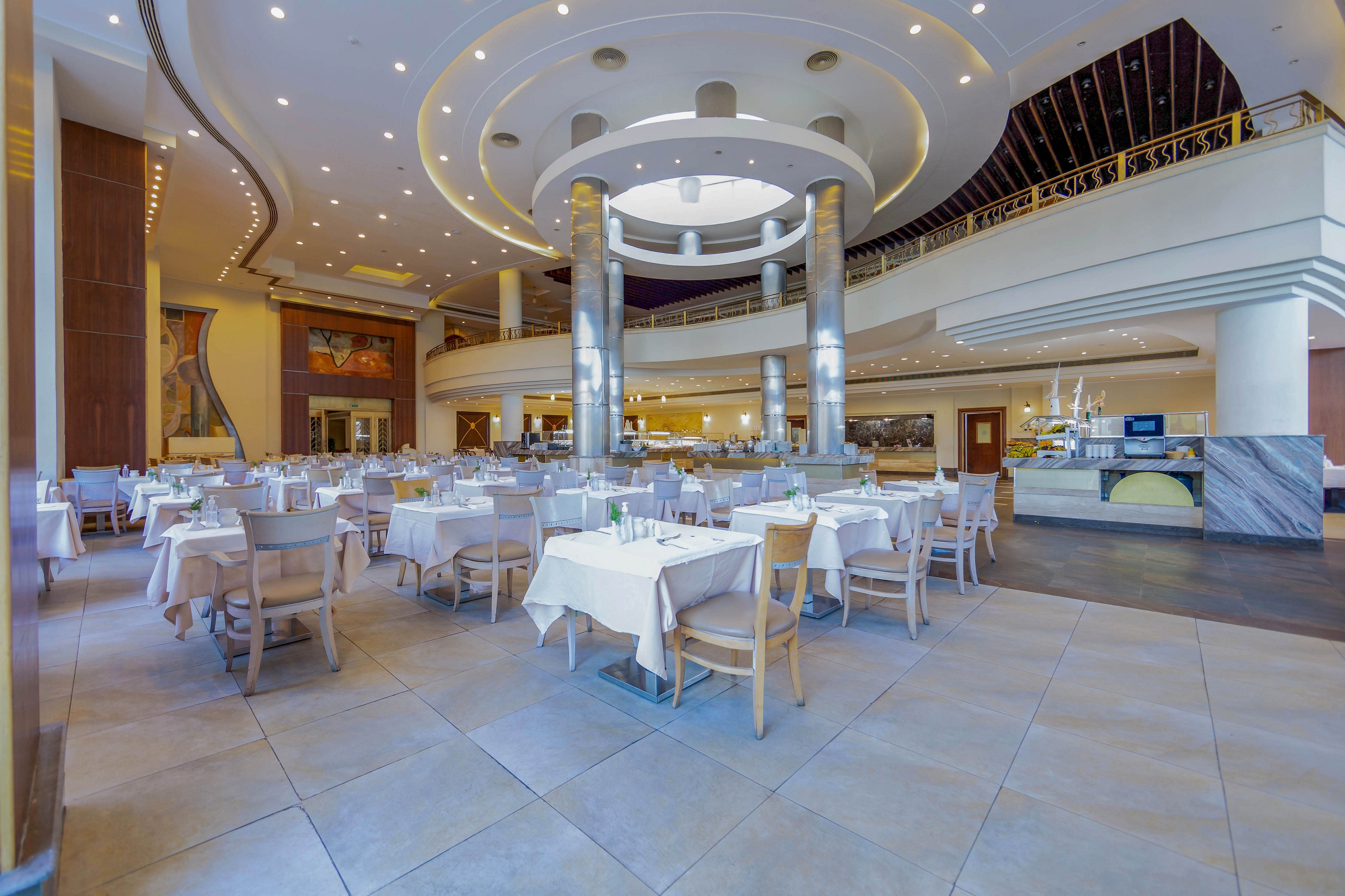 A bright and spacious restaurant interior featuring neatly arranged tables with white tablecloths and wooden chairs. The setting includes a high ceiling with elegant lighting fixtures and a contemporary design. The open layout highlights a clean and inviting atmosphere, ideal for dining or events.