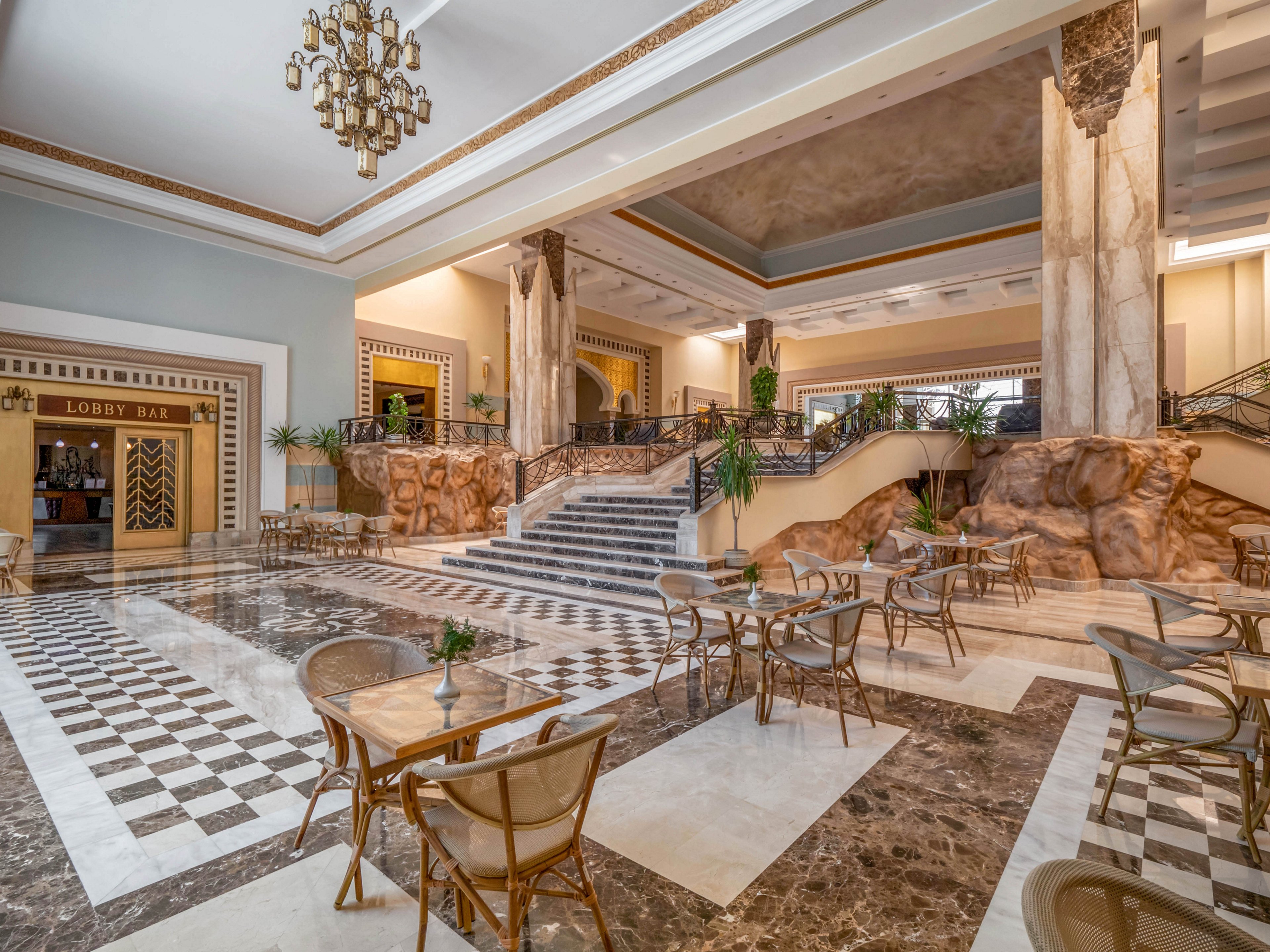 A spacious hotel lobby featuring polished marble flooring and a grand staircase. The area includes a seating arrangement with glass-top tables and chairs, surrounded by decorative plants. The 'Lobby Bar' is visible in the background, adding to the luxurious ambiance.