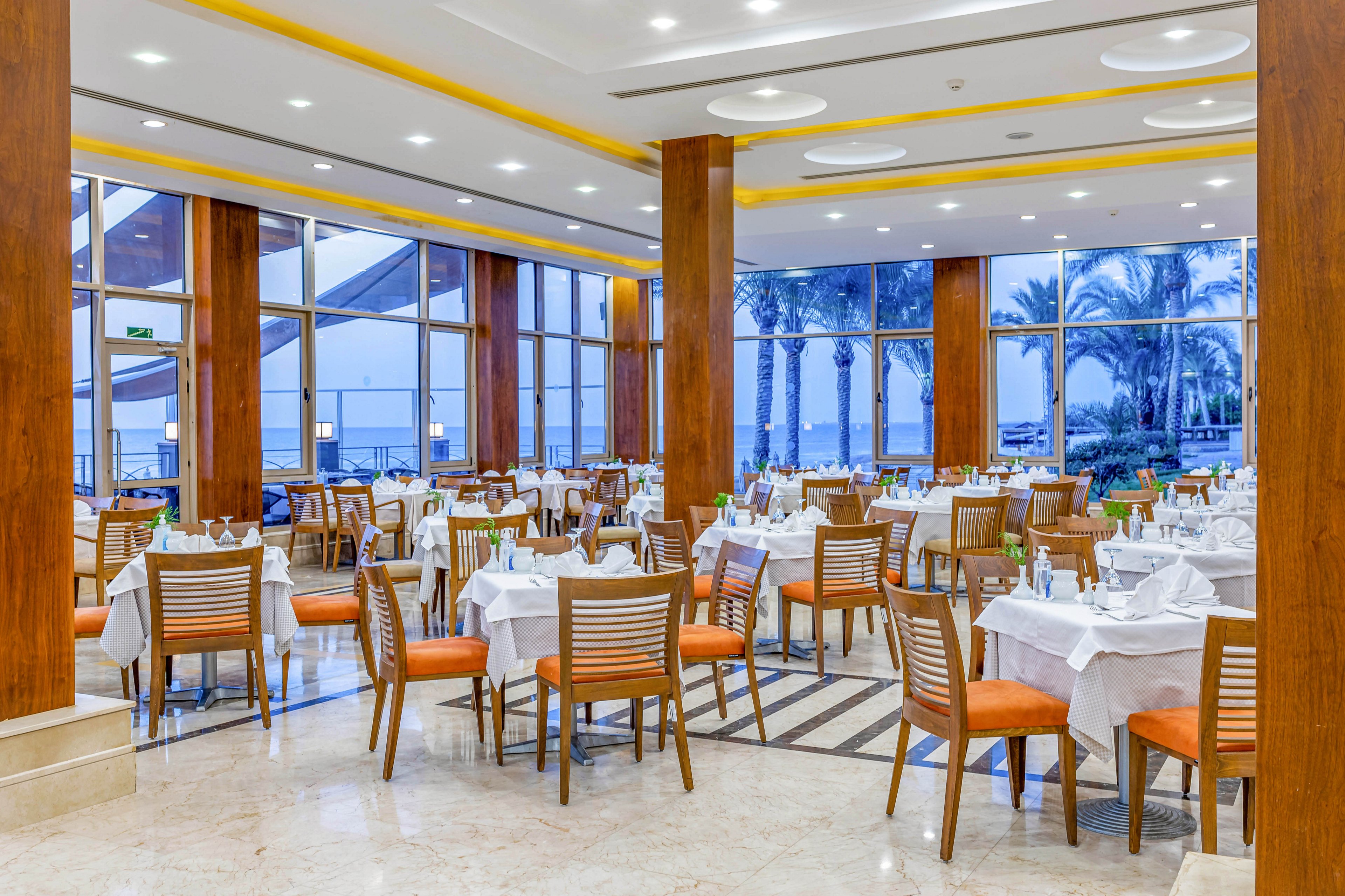 A spacious and well-lit restaurant featuring neatly arranged tables with white tablecloths and wooden chairs. Large glass windows provide a stunning view of palm trees and the ocean. The interior design includes warm wooden accents and modern lighting, creating a welcoming atmosphere.