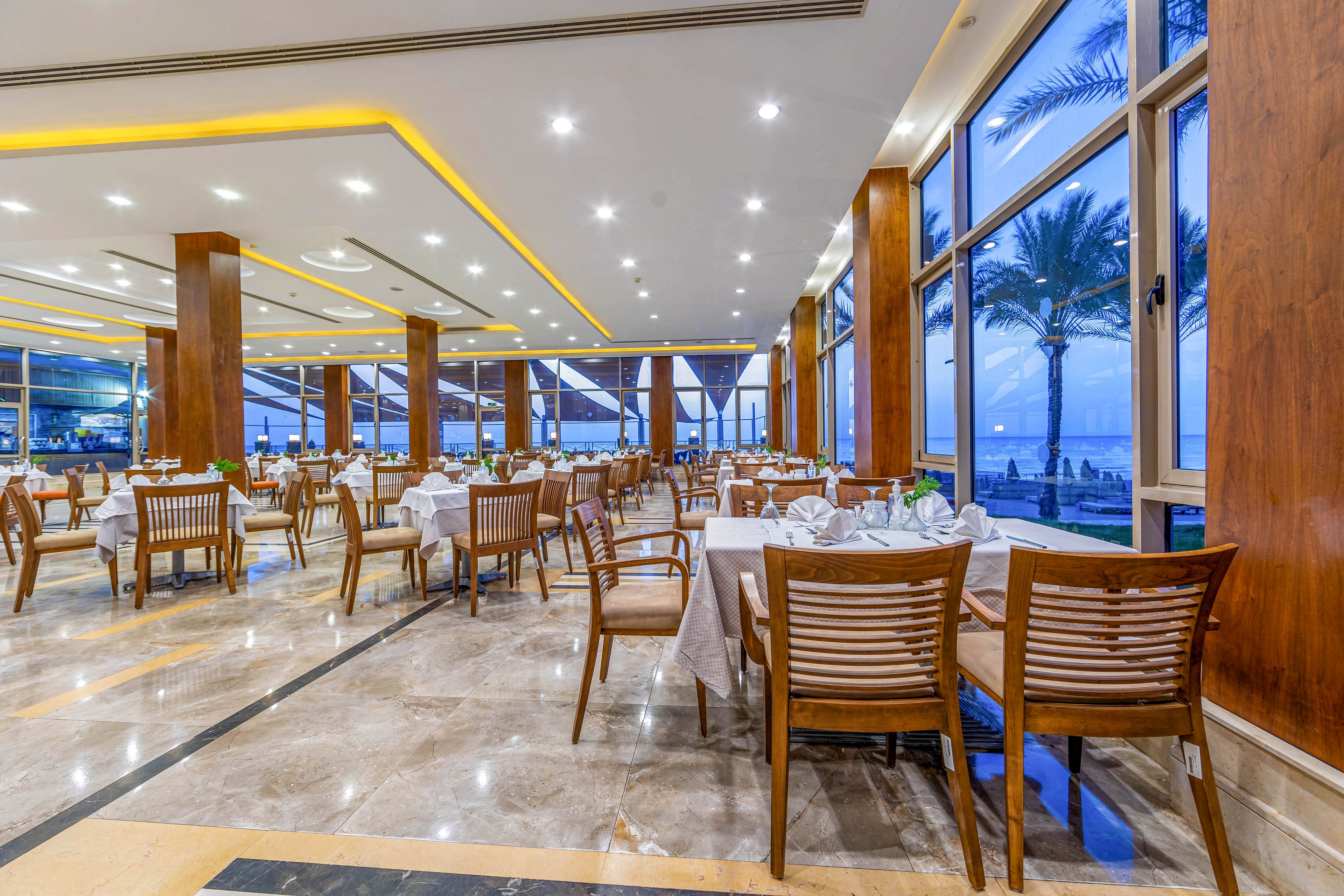 A spacious and well-lit restaurant featuring modern wooden furniture and large windows overlooking a seaside view. The interior is adorned with neatly arranged tables and chairs, creating a sophisticated dining atmosphere. The warm lighting complements the serene blue tones of the ocean visible through the windows.