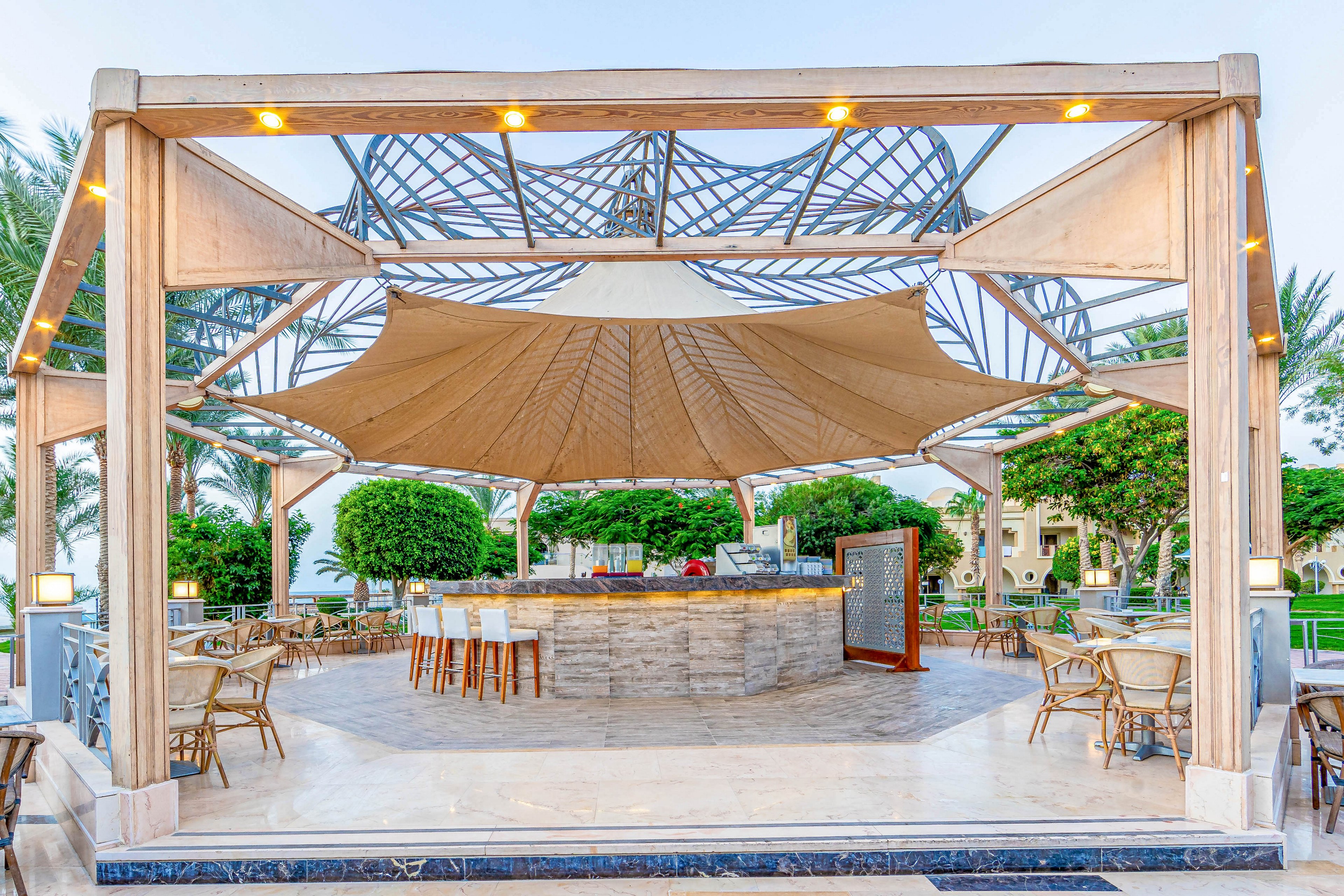 An elegant outdoor bar with a modern canopy structure, surrounded by bar stools and dining tables. The setting features lush greenery and a serene atmosphere, ideal for relaxation or social gatherings. The design incorporates a mix of wood and metal elements, creating a stylish and inviting space.