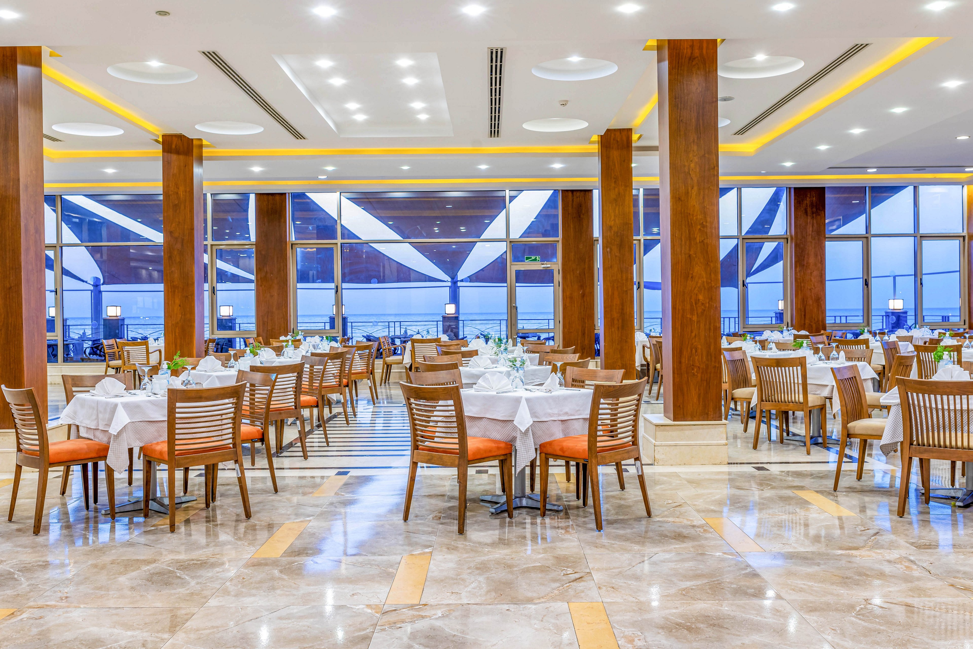 A spacious and well-lit restaurant featuring neatly arranged tables with white tablecloths and wooden chairs. The modern interior includes polished floors, wooden pillars, and large windows offering a view of the outdoors. The warm lighting and symmetrical layout create a welcoming and sophisticated atmosphere.