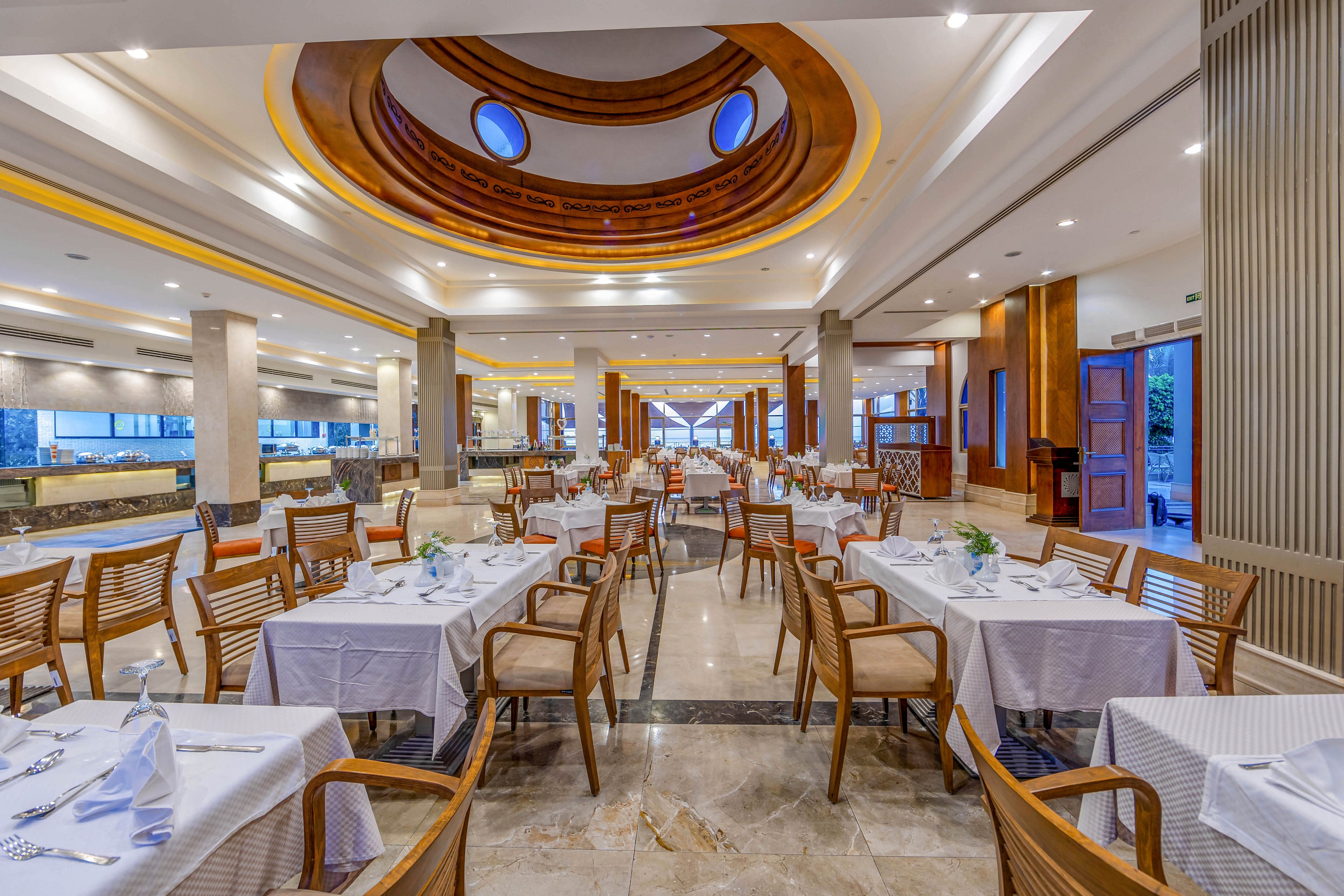 A spacious and well-lit restaurant interior featuring neatly arranged tables with white tablecloths and wooden chairs. The ceiling showcases a circular design with recessed lighting and skylights, adding a modern touch. The open layout and warm lighting create an inviting dining atmosphere.