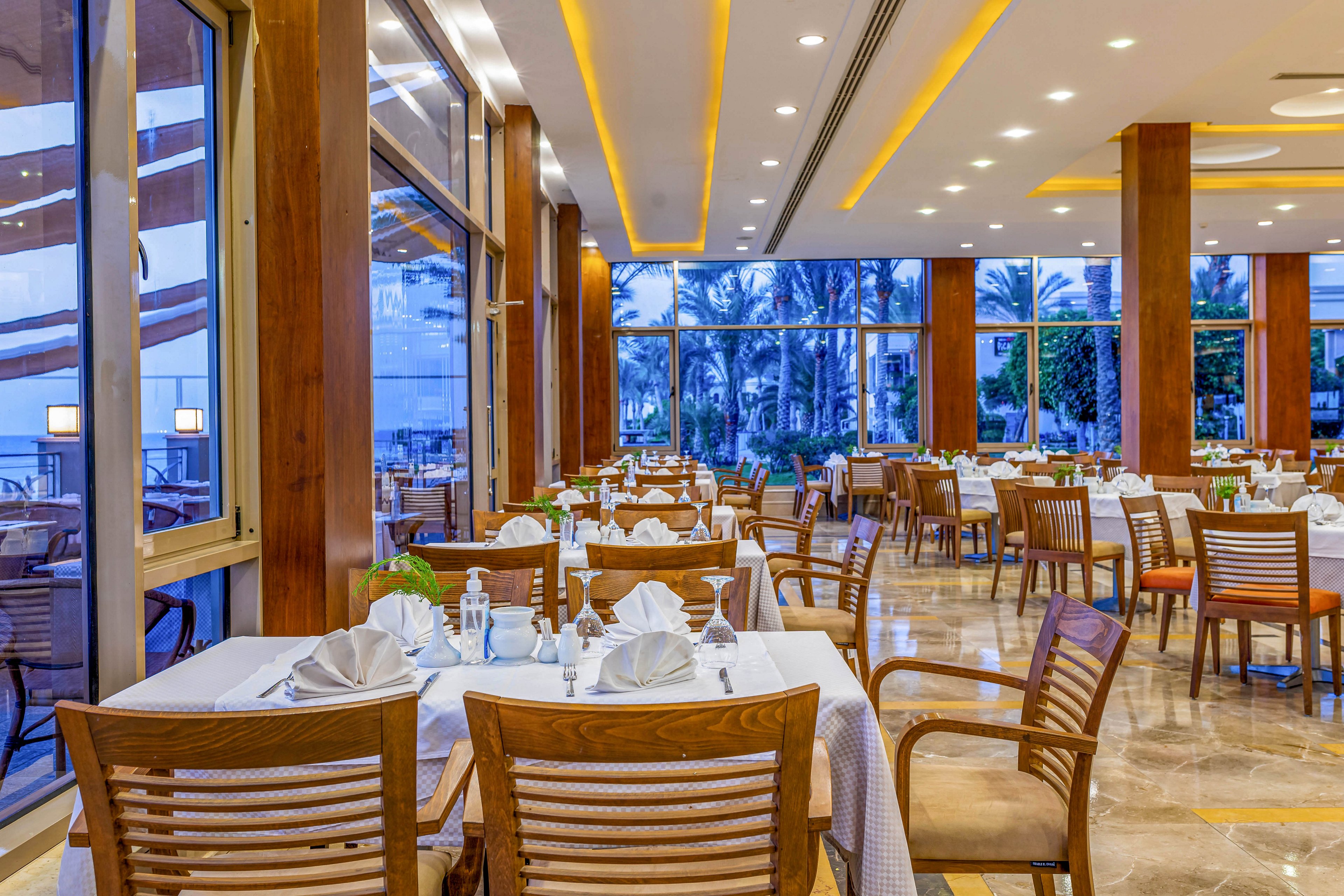 A spacious and well-lit restaurant featuring neatly arranged wooden tables and chairs, adorned with white tablecloths and folded napkins. Large glass windows provide a stunning view of palm trees and the ocean. The warm lighting and polished flooring create a welcoming and upscale dining atmosphere.