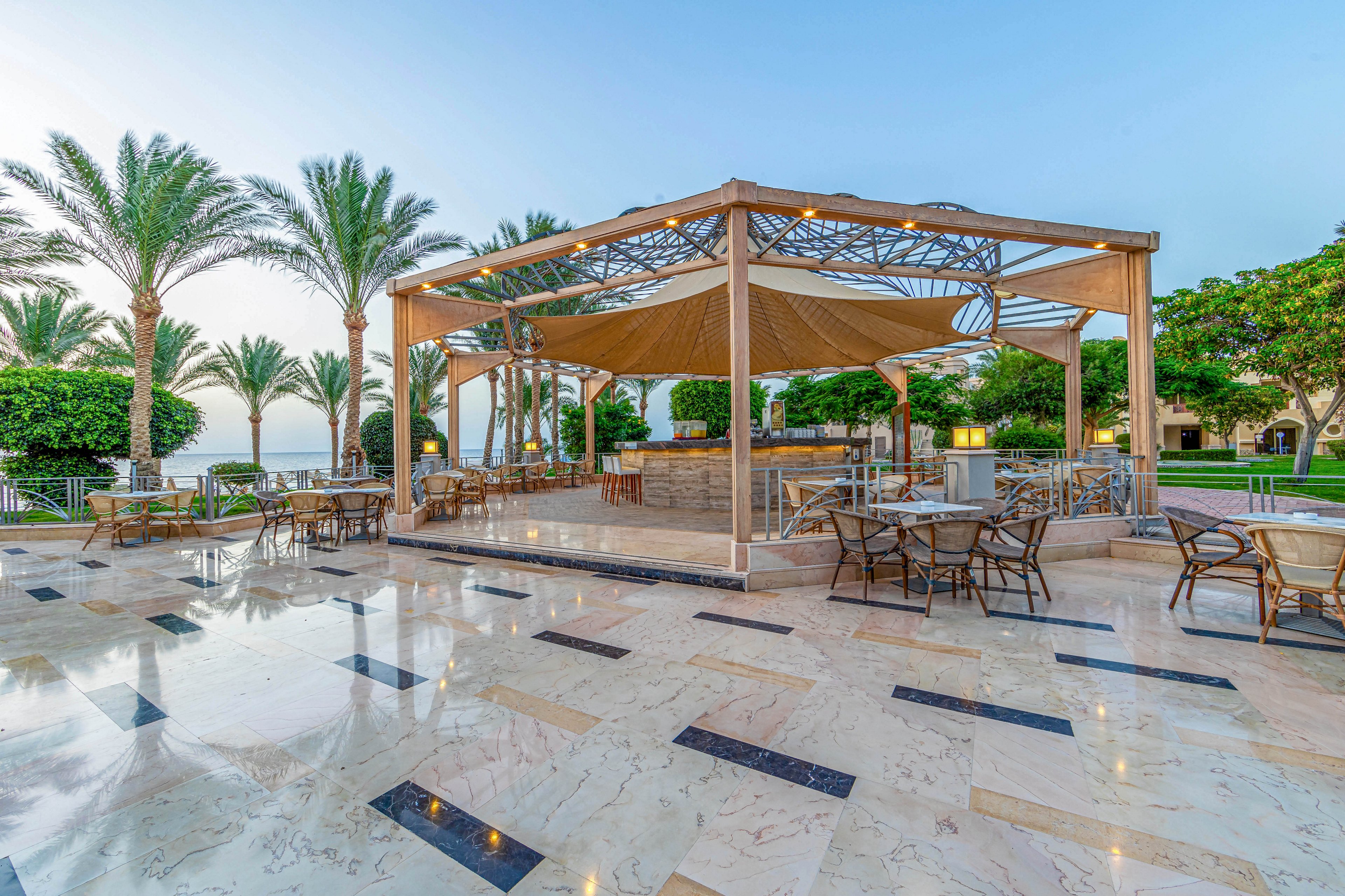 An elegant outdoor seating area featuring a modern canopy structure with metal and fabric elements. The setting includes marble flooring, scattered tables and chairs, and lush greenery with palm trees in the background. The scene is illuminated by soft lighting, creating a serene and inviting atmosphere.