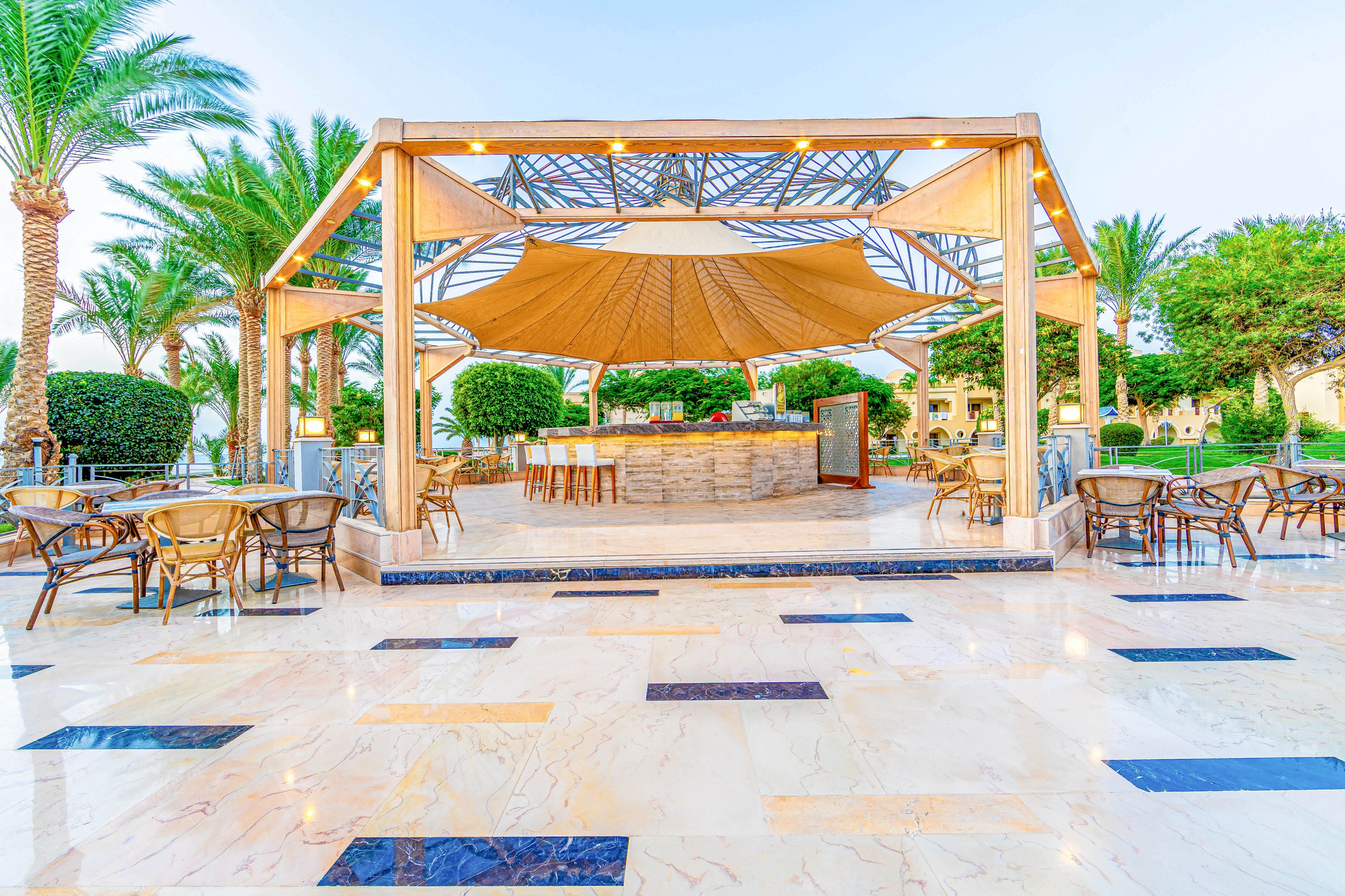 An inviting outdoor cafe with a central bar area under a large canopy structure. Surrounded by palm trees and greenery, the setting features multiple tables and chairs on a polished tiled floor. The bright and open atmosphere is enhanced by warm lighting and a clear sky.