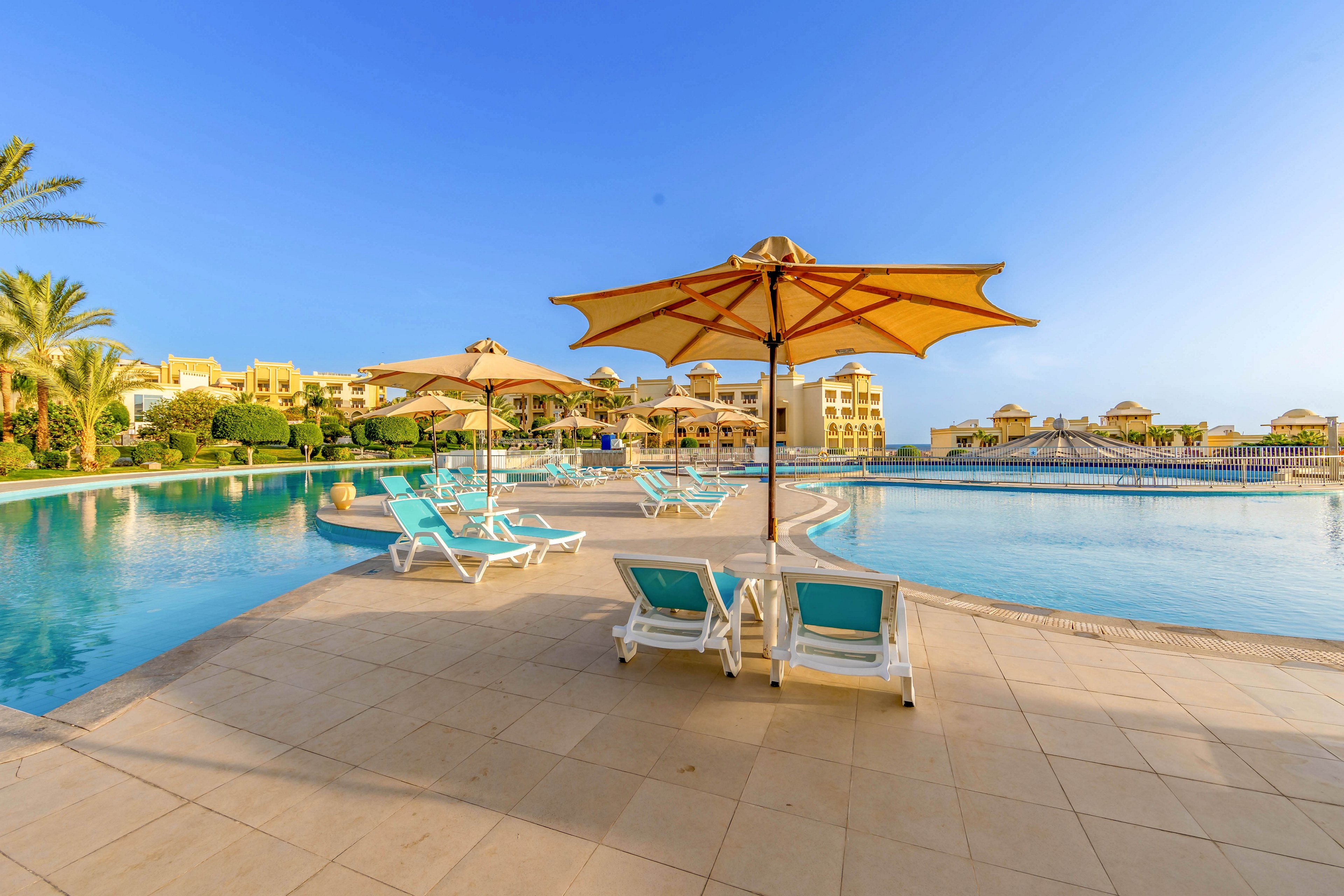 A large, inviting swimming pool is surrounded by sun loungers and umbrellas, set against a backdrop of resort buildings. The scene is tranquil, with clear blue skies and palm trees adding to the vacation atmosphere. The pool deck is spacious, and the water reflects the bright sunlight, creating a relaxing mood. No people are present, emphasizing a peaceful and serene setting.