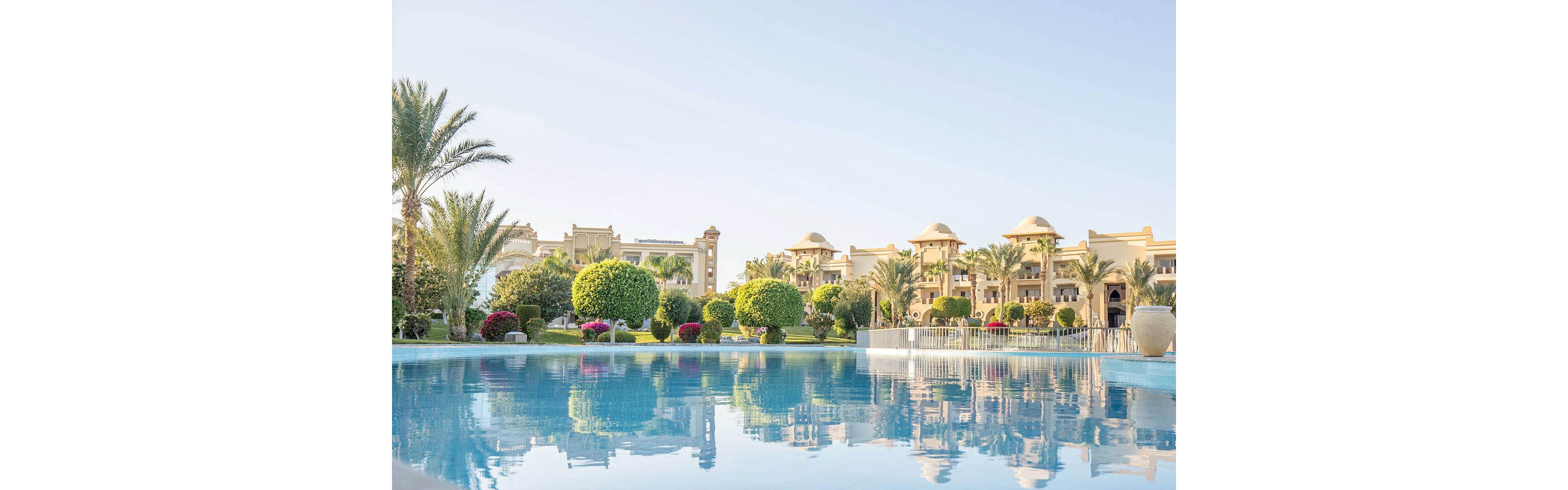 A serene luxury resort is shown with a large, clear swimming pool in the foreground. The setting features manicured gardens, palm trees, and elegant buildings with domed rooftops under a clear blue sky. The tranquil atmosphere is enhanced by the reflection of the architecture and greenery in the pool. No visible text or numbers are present in the image.