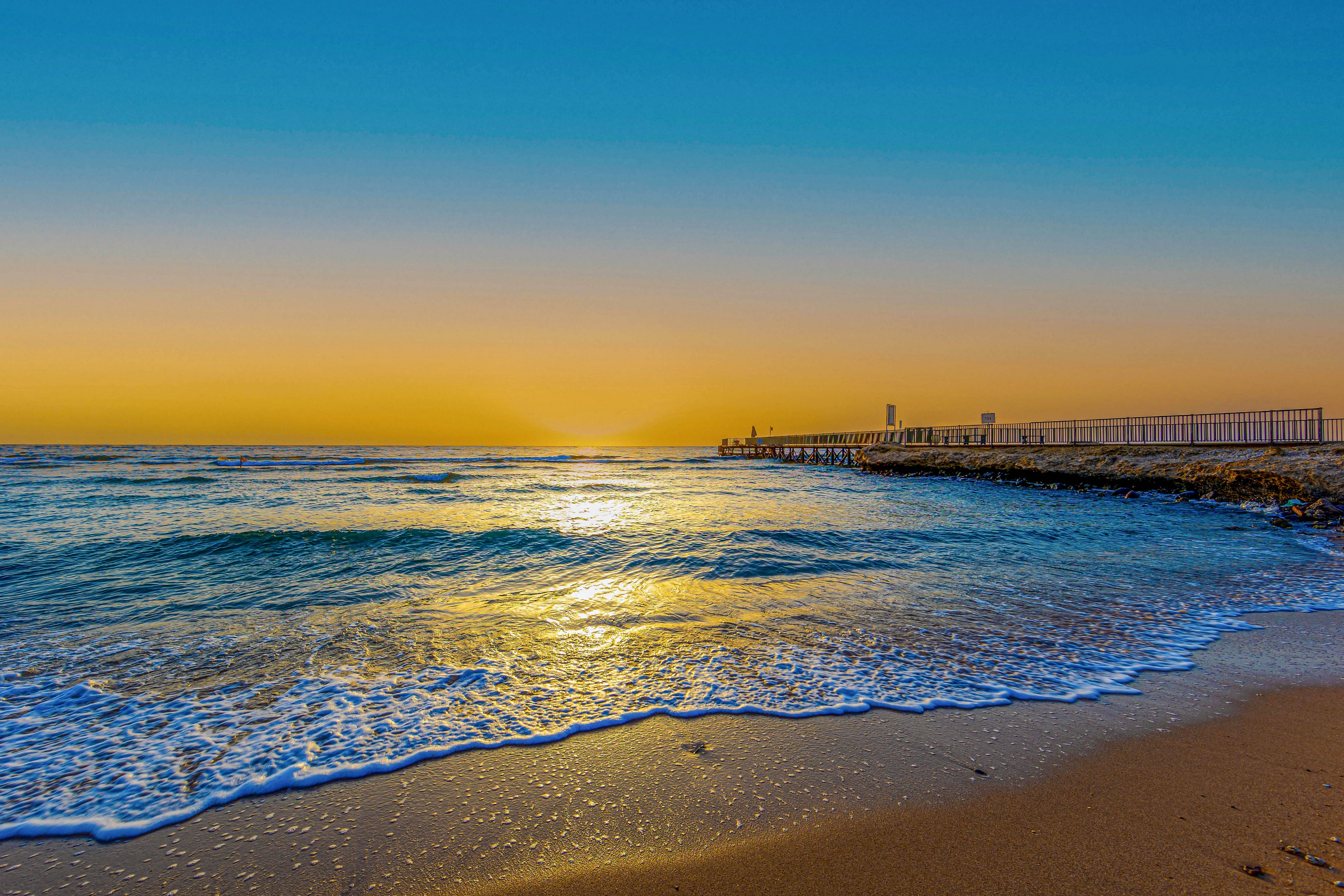 A serene sunrise casts golden hues across a calm ocean, gently lapping onto a sandy beach. A long pier extends into the water on the right, silhouetted against the vibrant sky. The scene features clear blue and yellow tones, evoking a peaceful and refreshing mood. No people or visible text are present in the image.