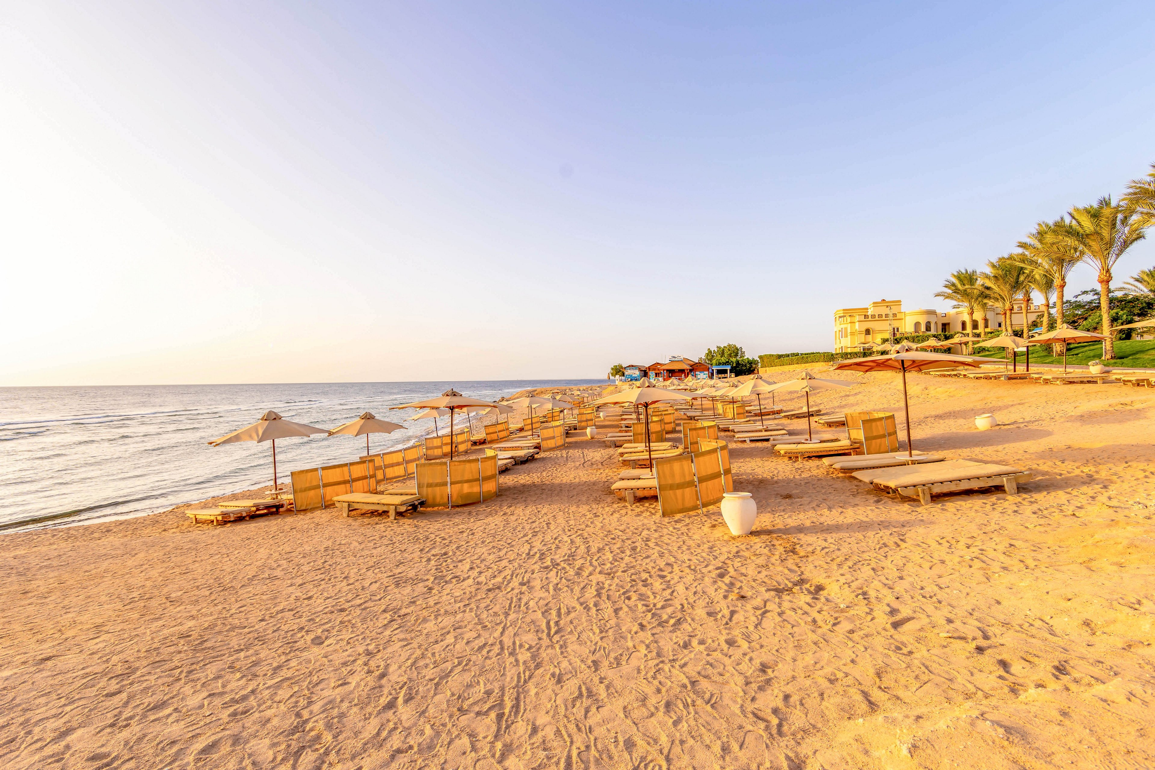 A tranquil sandy beach is lined with empty wooden sun loungers and straw umbrellas, facing the calm sea under a clear sky. The early morning sunlight casts a warm golden glow over the scene, with a resort building and palm trees visible in the background. The beach appears peaceful and inviting, ideal for relaxation and leisure.