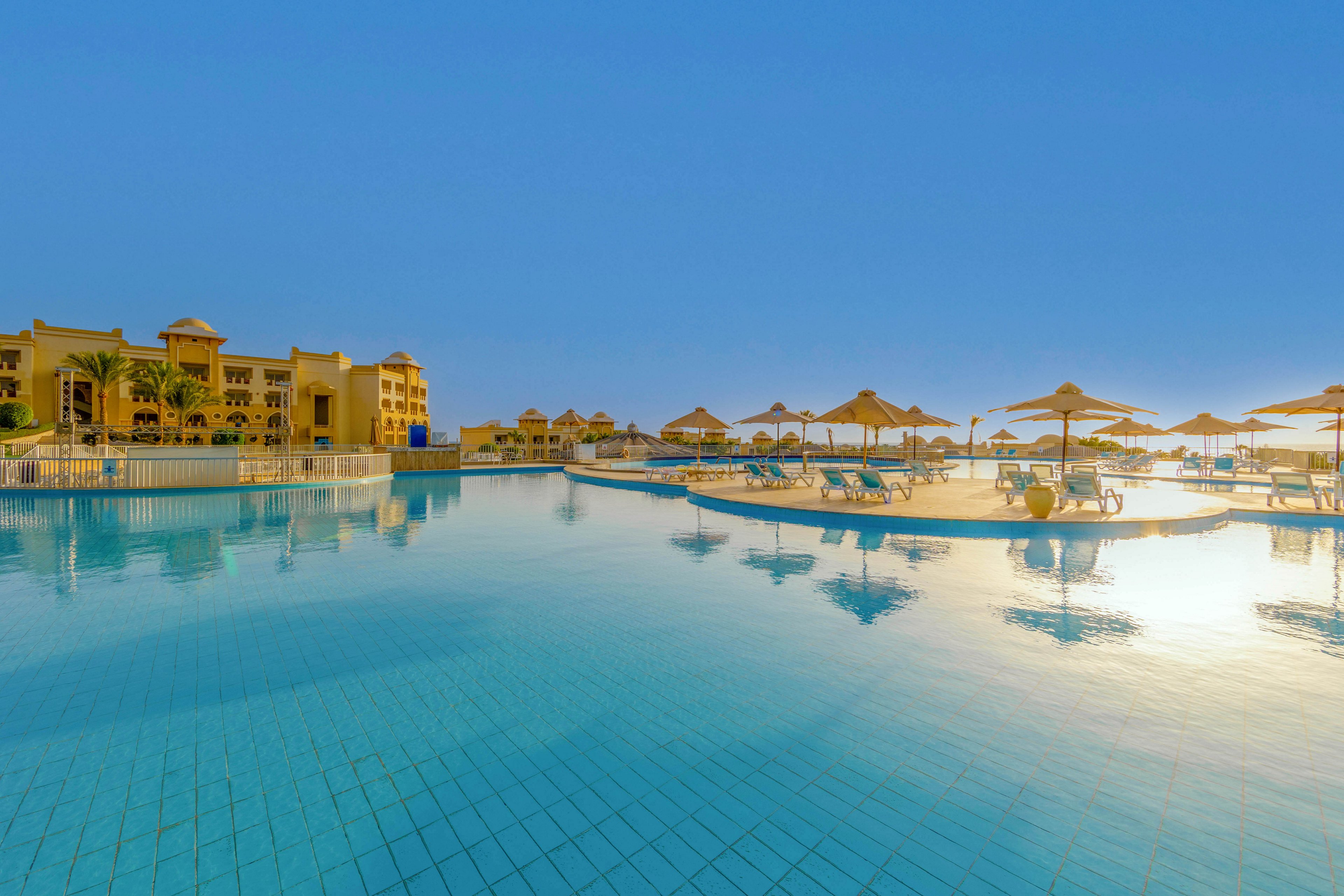 A large, tranquil swimming pool is the central focus, surrounded by sun loungers and umbrellas. Elegant resort buildings are visible in the background, set against a clear blue sky. The scene evokes a relaxing, vacation atmosphere with warm, golden sunlight reflecting off the water. No visible text or numbers are present in the image.