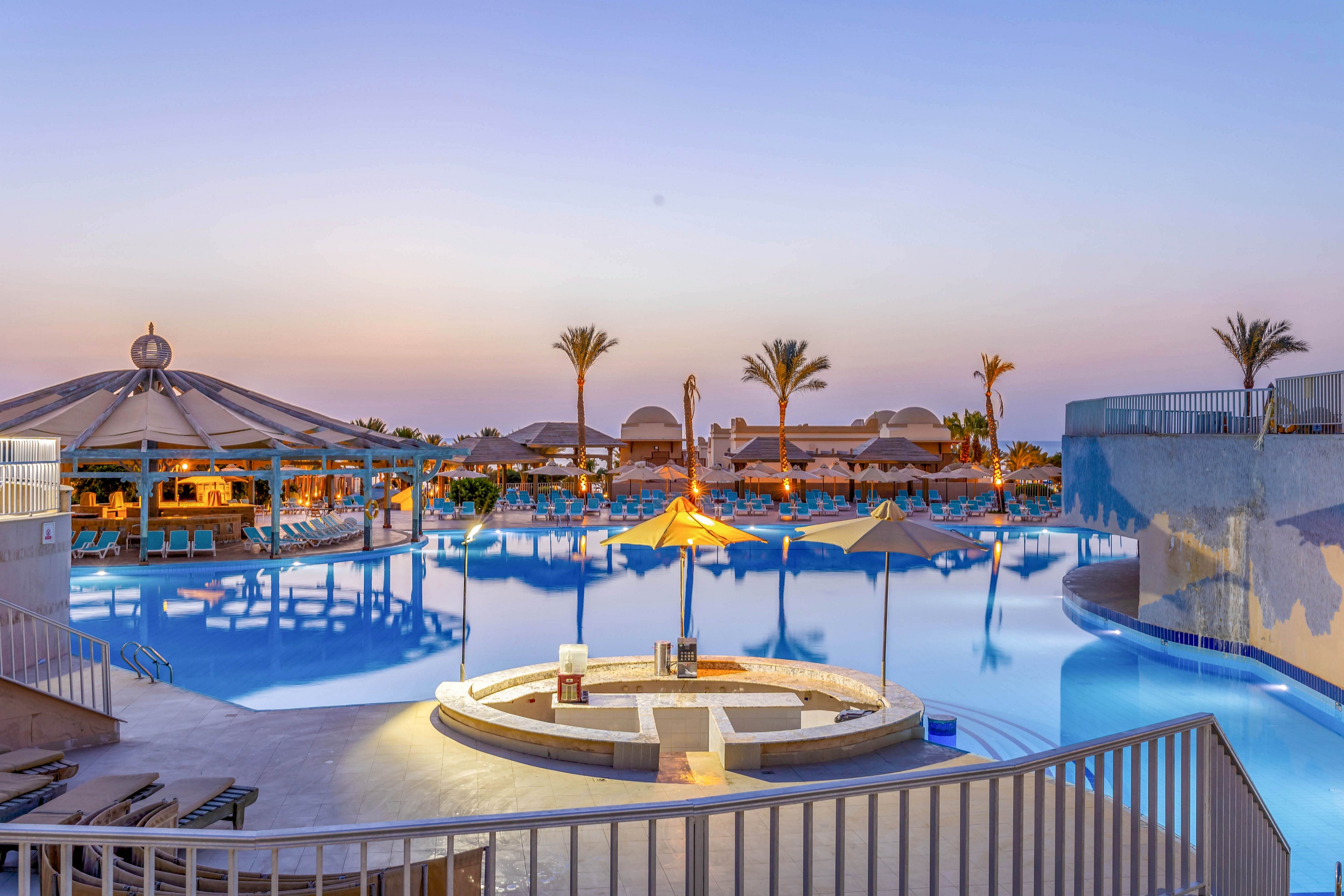A tranquil resort pool is featured at dusk, surrounded by palm trees and modern lounge areas. The setting includes sun umbrellas, a circular seating area, and softly illuminated water reflecting the evening sky. The atmosphere is calm and inviting, with warm lighting and clear architectural lines. No visible text or numbers are present in the image.