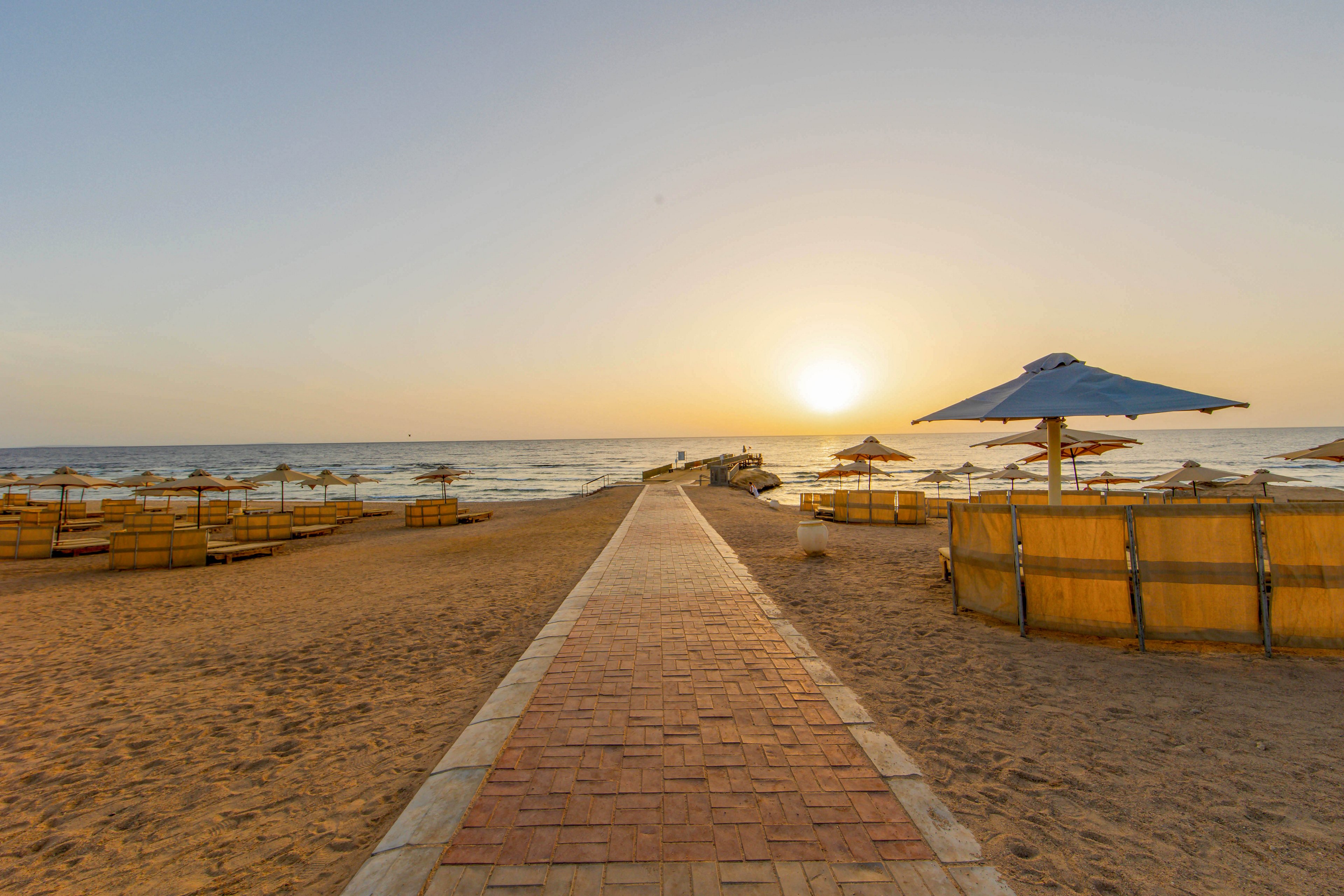 A paved walkway leads directly to the shoreline, flanked by straw beach umbrellas and wooden cabanas on a sandy beach. The sun is setting over the calm sea, casting a warm golden light across the scene. The beach appears empty, evoking a tranquil and serene atmosphere.