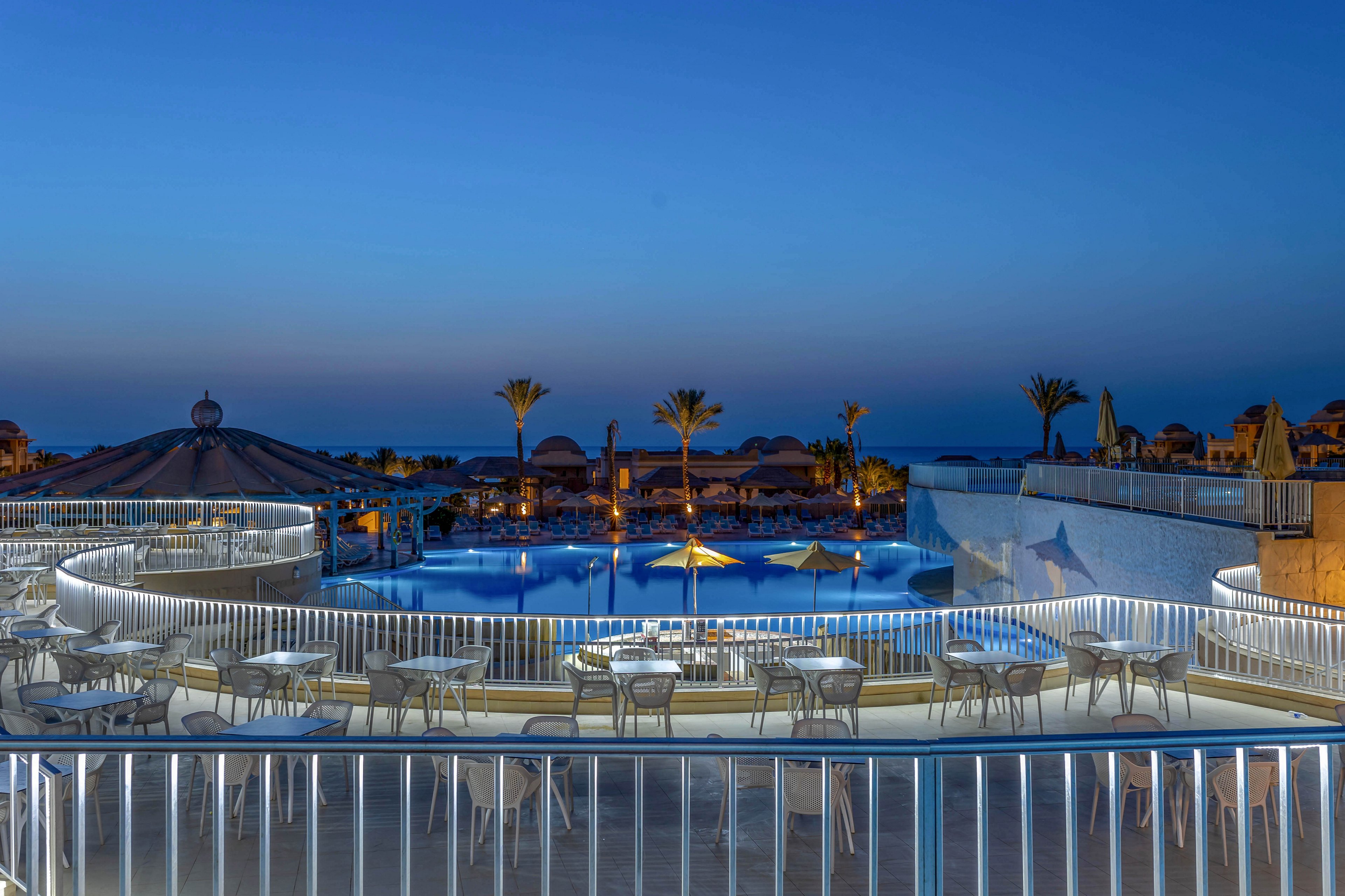 A tranquil outdoor swimming pool is surrounded by a terrace with white tables and chairs, illuminated by soft lighting at dusk. Palm trees and thatched umbrellas line the pool area, creating a relaxing vacation atmosphere. The sky transitions from deep blue to twilight, and the calm water reflects the ambient lights. No visible text or numbers are present in the image.