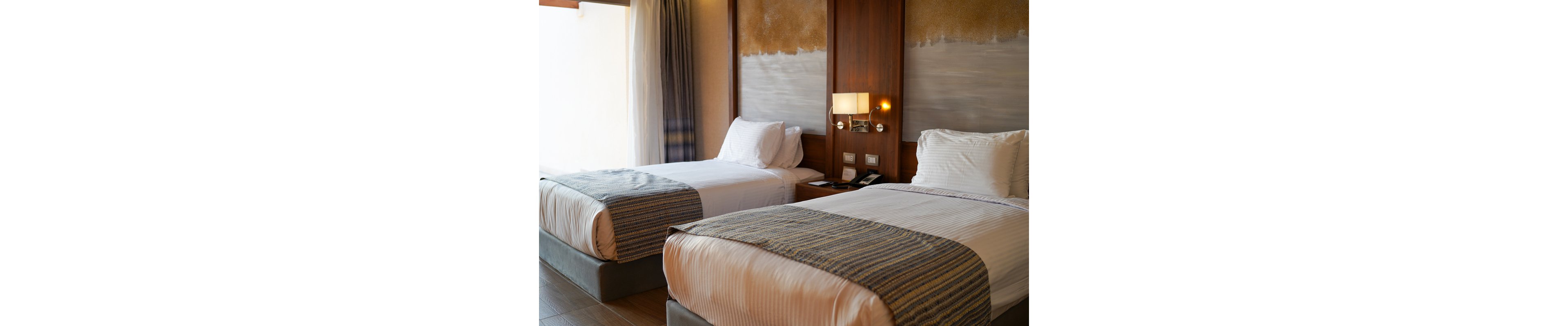 A cozy hotel room featuring two neatly made twin beds with striped bed runners. The room has a warm wooden headboard, soft lighting from wall-mounted lamps, and a neutral color palette. A bedside table with a phone and power outlets adds functionality to the space.