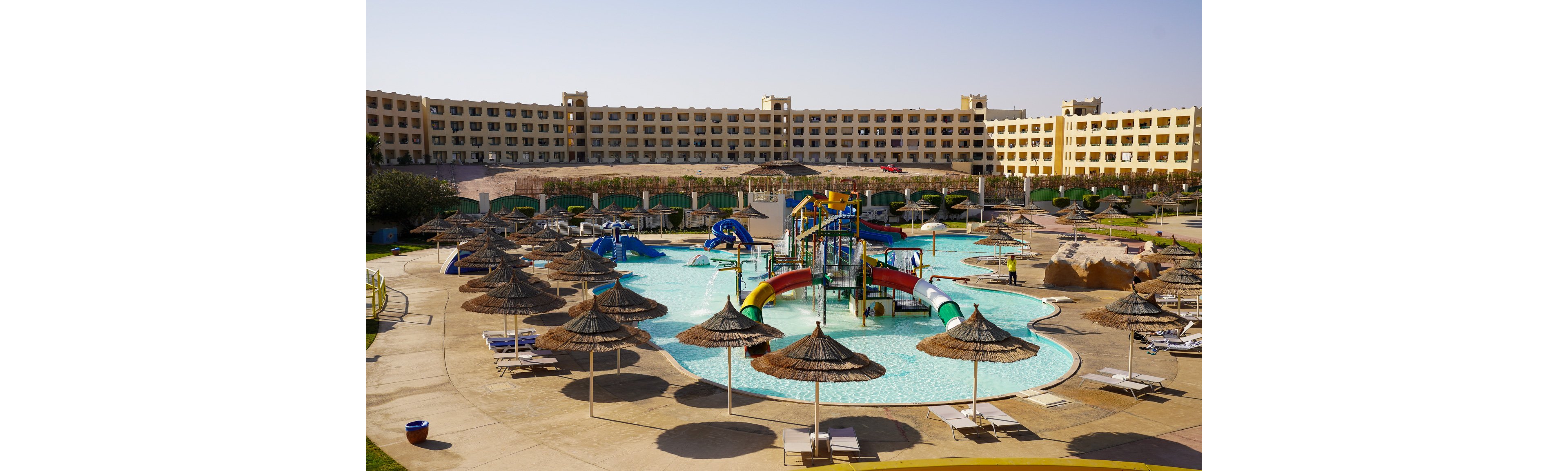 Large resort-style swimming pool features a central colorful water playground with slides and climbing structures. Numerous straw parasols and sun loungers surround the pool, suggesting a relaxing vacation setting. A multi-story hotel building forms the backdrop under a clear sky, emphasizing a warm, sunny climate. The scene appears calm with no visible people, highlighting the facilities and architecture.