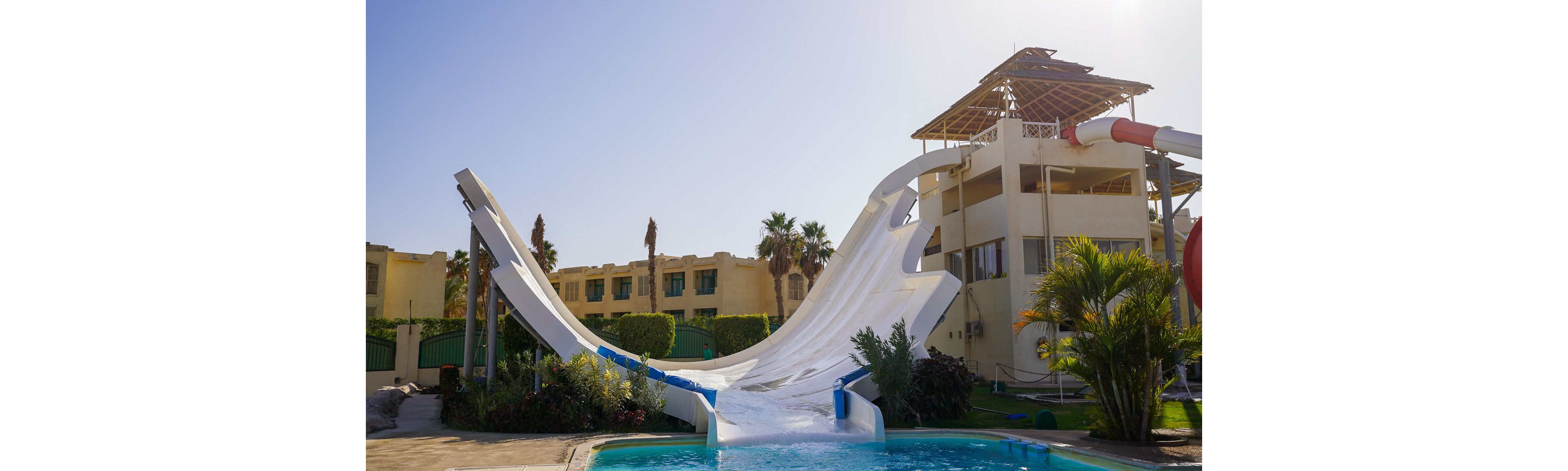 A tall, white water slide with steep curved ramps descends into a clear blue pool at an outdoor resort. The slide structure is attached to a multi-story building with open balconies and a partially covered rooftop. Palm trees, shrubs, and other greenery surround the pool area, adding a tropical feel. In the background, additional resort buildings and a bright blue sky suggest a warm, sunny vacation setting.