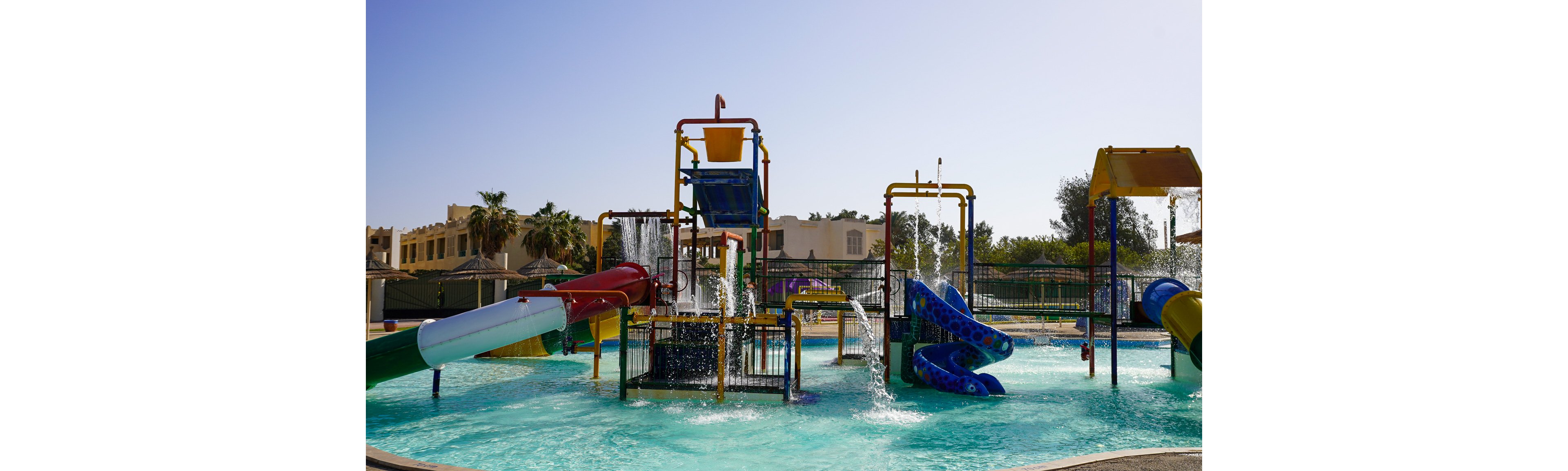 A vibrant outdoor water playground features multiple colorful slides and splash structures in a shallow pool. Water pours from overhead buckets and pipes, creating fountains and streams across the play area. The scene is set under a clear blue sky with buildings, trees, and palm trees in the background. The bright colors and sunny weather suggest a fun, family-friendly recreational environment.
