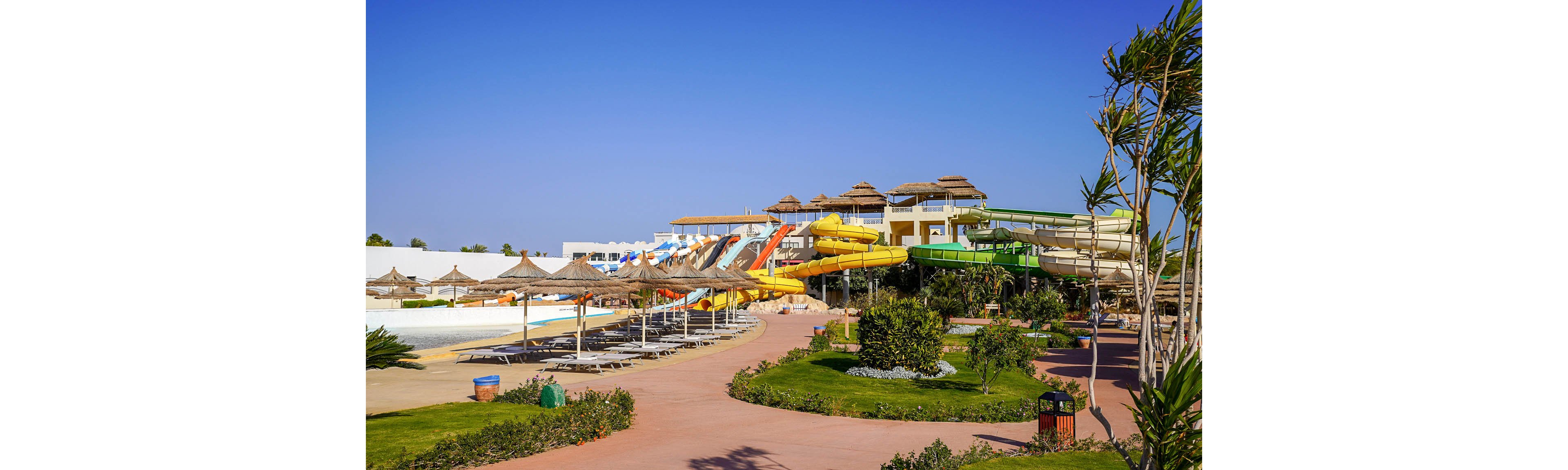 A large outdoor water park features multiple colorful slides, including orange, yellow, and green tubes, attached to a multi-level structure. In the foreground, sun loungers and straw parasols line a wide pathway and poolside area. Lush green shrubs and palm-like plants add a tropical feel to the scene. The bright blue sky and strong sunlight create a warm, inviting vacation atmosphere.