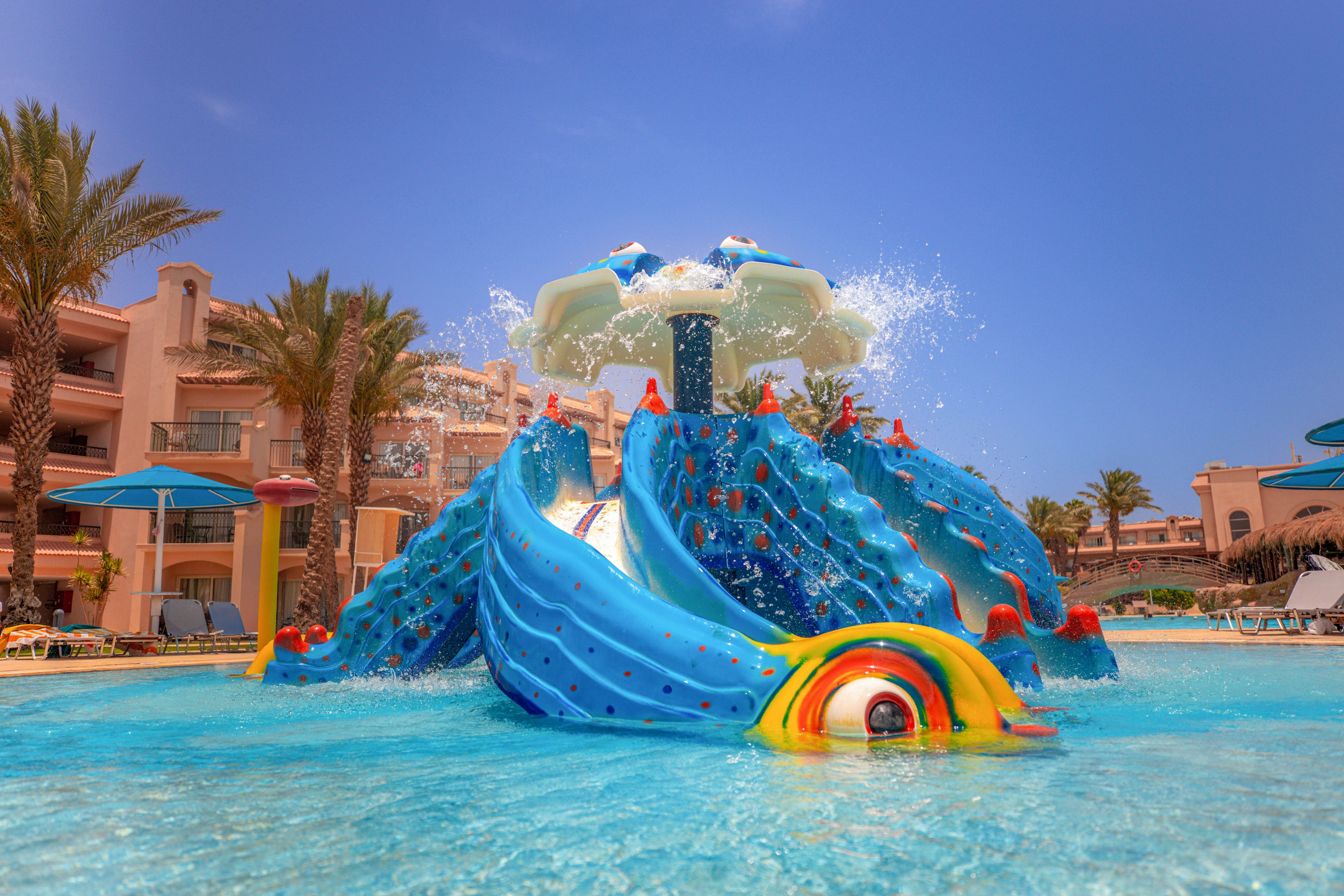 A vibrant water playground features large blue slides shaped like a sea creature, set in a shallow pool. Water sprays from the top structure, creating a lively and refreshing atmosphere under a clear blue sky. Palm trees and resort buildings surround the play area, enhancing the vacation vibe. The scene is bright and inviting, ideal for children and families enjoying a sunny day.