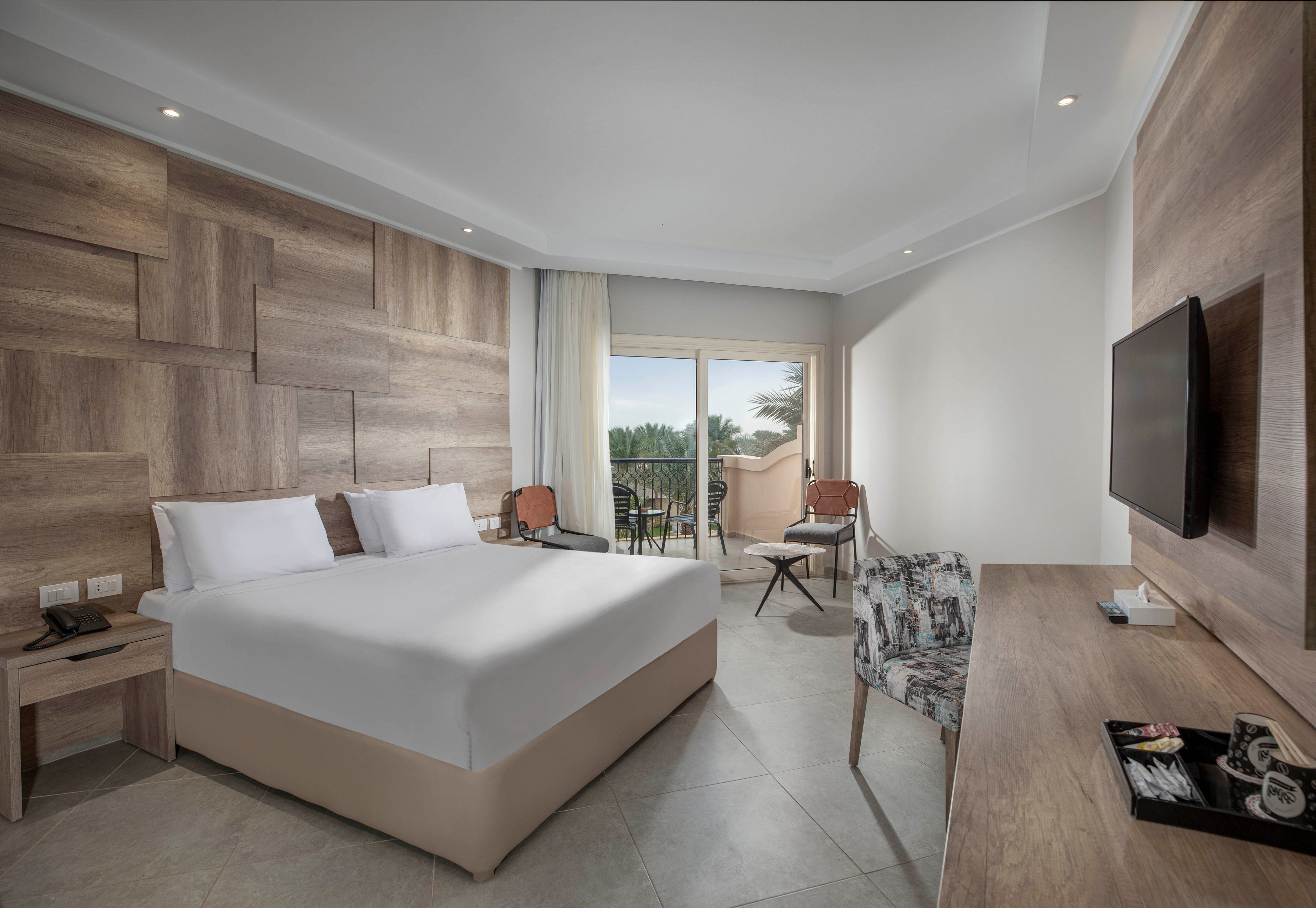 A spacious hotel room features a large bed with crisp white linens, a wooden accent wall, and a sleek desk area. The room opens to a balcony with outdoor seating, offering a view of palm trees and natural light. Neutral tones and contemporary furnishings create a calm, inviting atmosphere, with amenities visible on the desk and bedside tables.