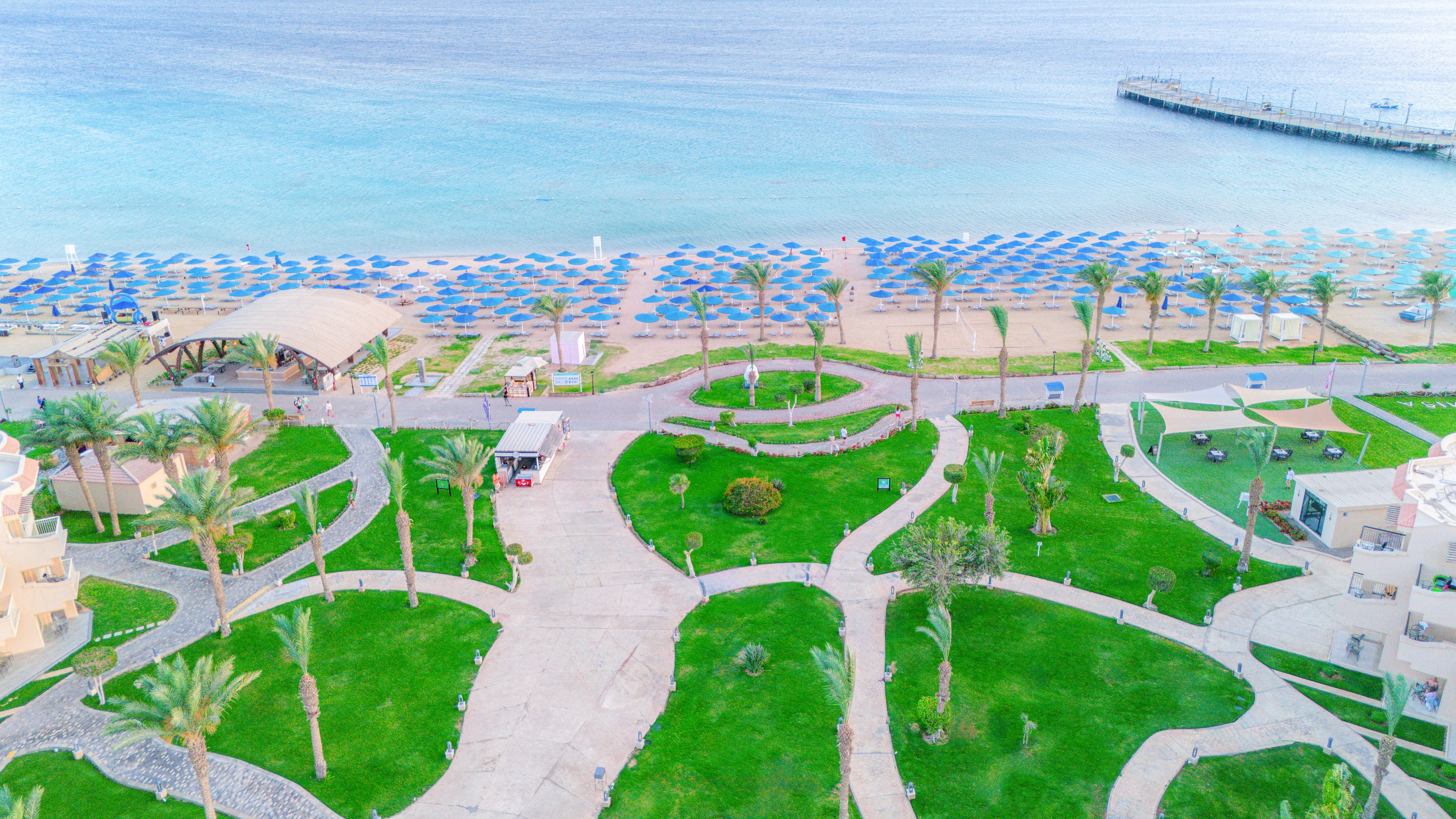 An aerial photograph captures a beachfront resort with lush green lawns and winding pathways leading to a sandy beach. Numerous blue umbrellas are neatly arranged along the shoreline, facing the calm, turquoise sea. The scene includes palm trees, a pier extending into the water, and resort buildings, creating a tranquil and inviting atmosphere. The overall palette is bright and tropical, emphasizing relaxation and leisure.