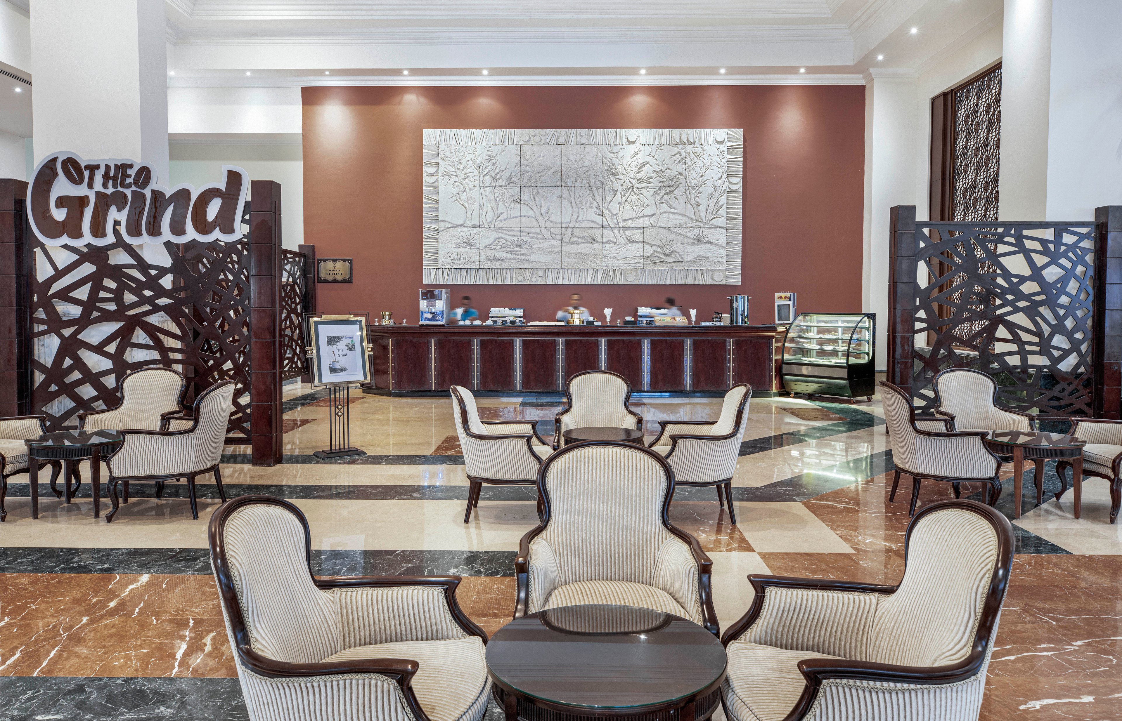 A stylish hotel café named 'The Grind' features plush armchairs and small tables arranged in a spacious, well-lit lounge. The setting includes a marble floor, decorative wooden partitions, and a large wall art piece behind the counter. The café counter displays various items and a glass case, with the name 'The Grind' clearly visible. The overall atmosphere is modern and inviting, suitable for relaxation or casual meetings.