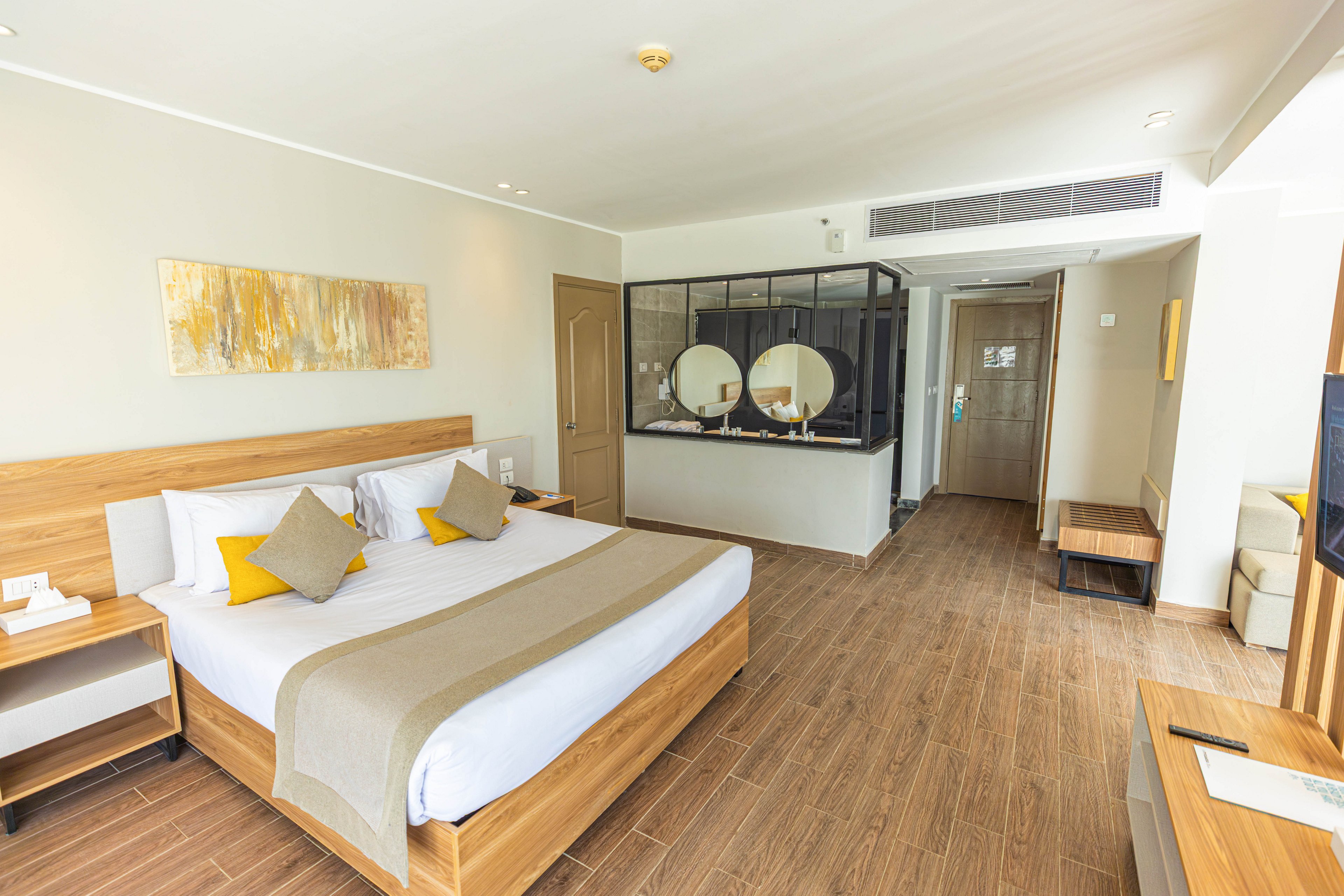 A spacious hotel room features a large king-sized bed with white linens and beige accents. The setting includes contemporary wooden furniture, a glass-walled bathroom area with dual sinks, and warm, neutral decor. Natural light fills the room, highlighting the clean lines and minimalist style. Decorative pillows and abstract wall art add a touch of color.