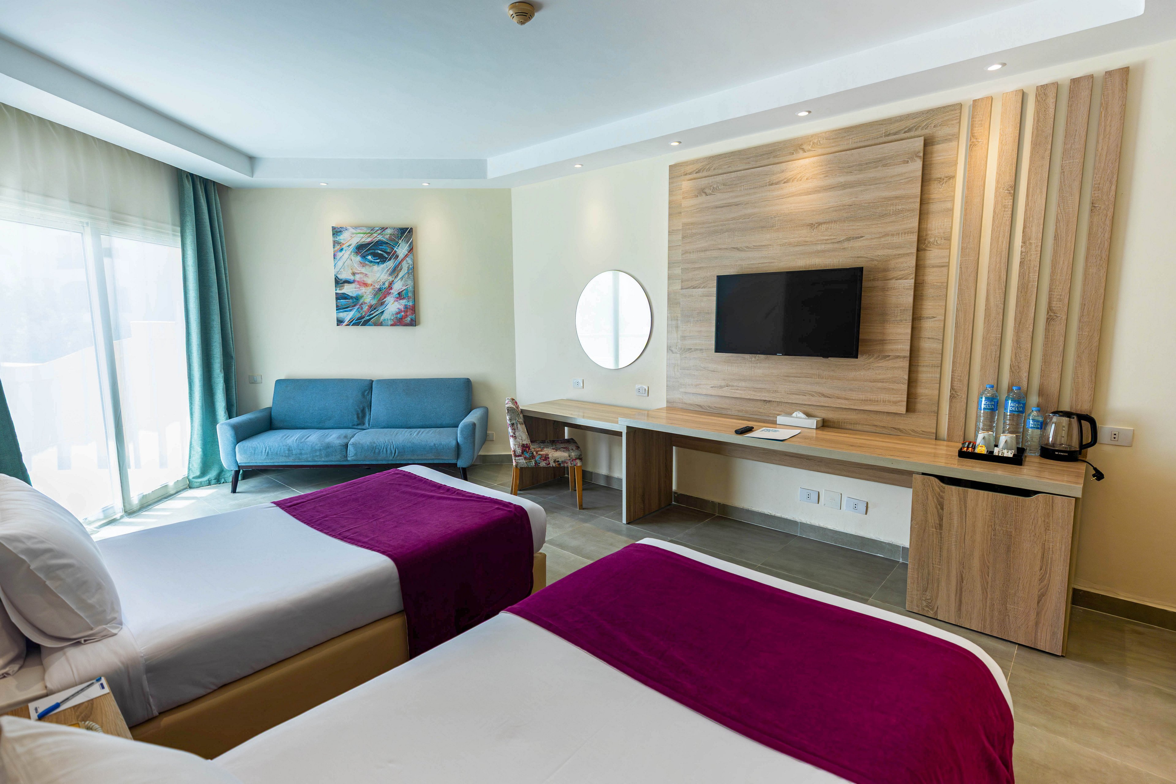 A contemporary hotel room features two twin beds with burgundy throws, a blue sofa, and a large window with sheer curtains. The workspace includes a wall-mounted TV, desk, and complimentary water bottles with a kettle. The room is decorated in neutral tones with a pop of color from the artwork and bedding, creating a clean and inviting atmosphere.