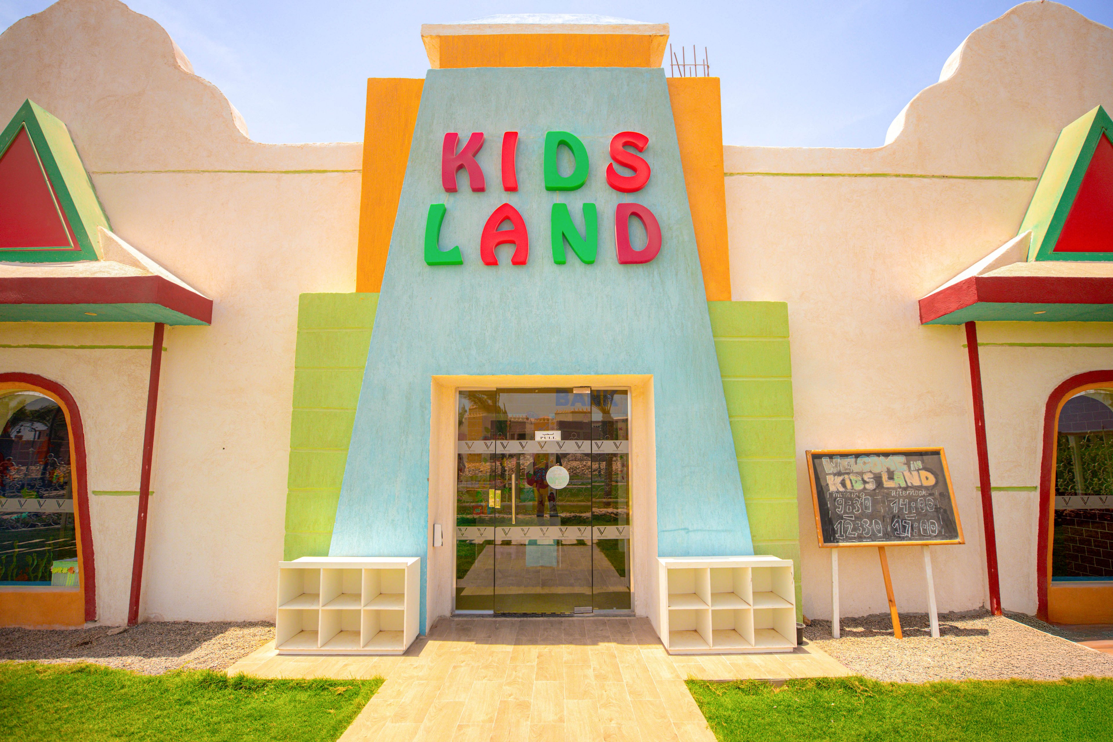 The image shows the entrance to a children's facility named 'KIDS LAND,' with large, colorful letters above the door. The building features a playful, geometric facade in pastel tones, and a chalkboard sign is visible to the right of the entrance. The setting is bright and sunny, suggesting a cheerful and welcoming atmosphere for families and children.