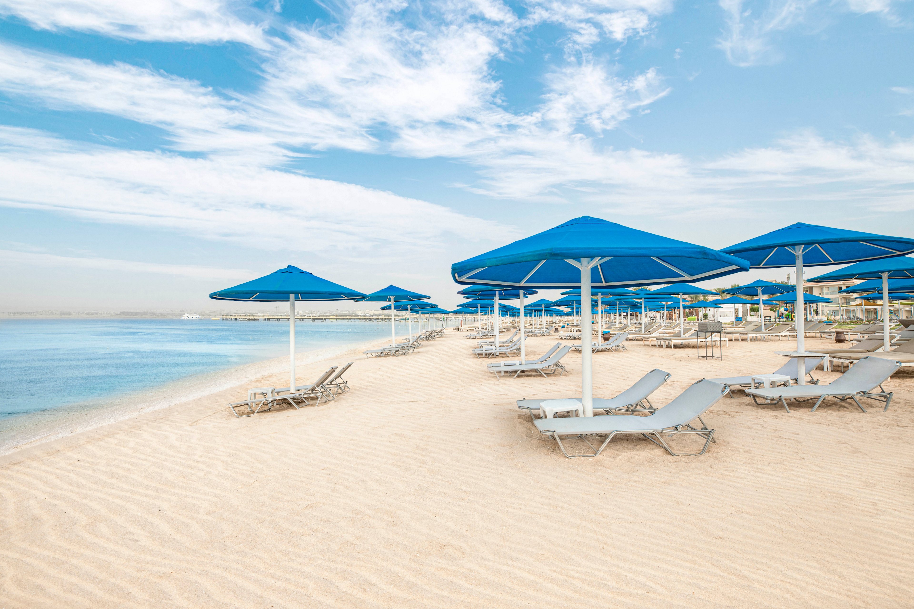A tranquil sandy beach features rows of blue sun umbrellas and empty lounge chairs facing a calm, clear shoreline. The setting is peaceful with soft, natural light and a few wispy clouds in the sky. The scene suggests a relaxing vacation destination, with no people present and a clean, inviting atmosphere. The overall palette is dominated by shades of blue, beige, and white.