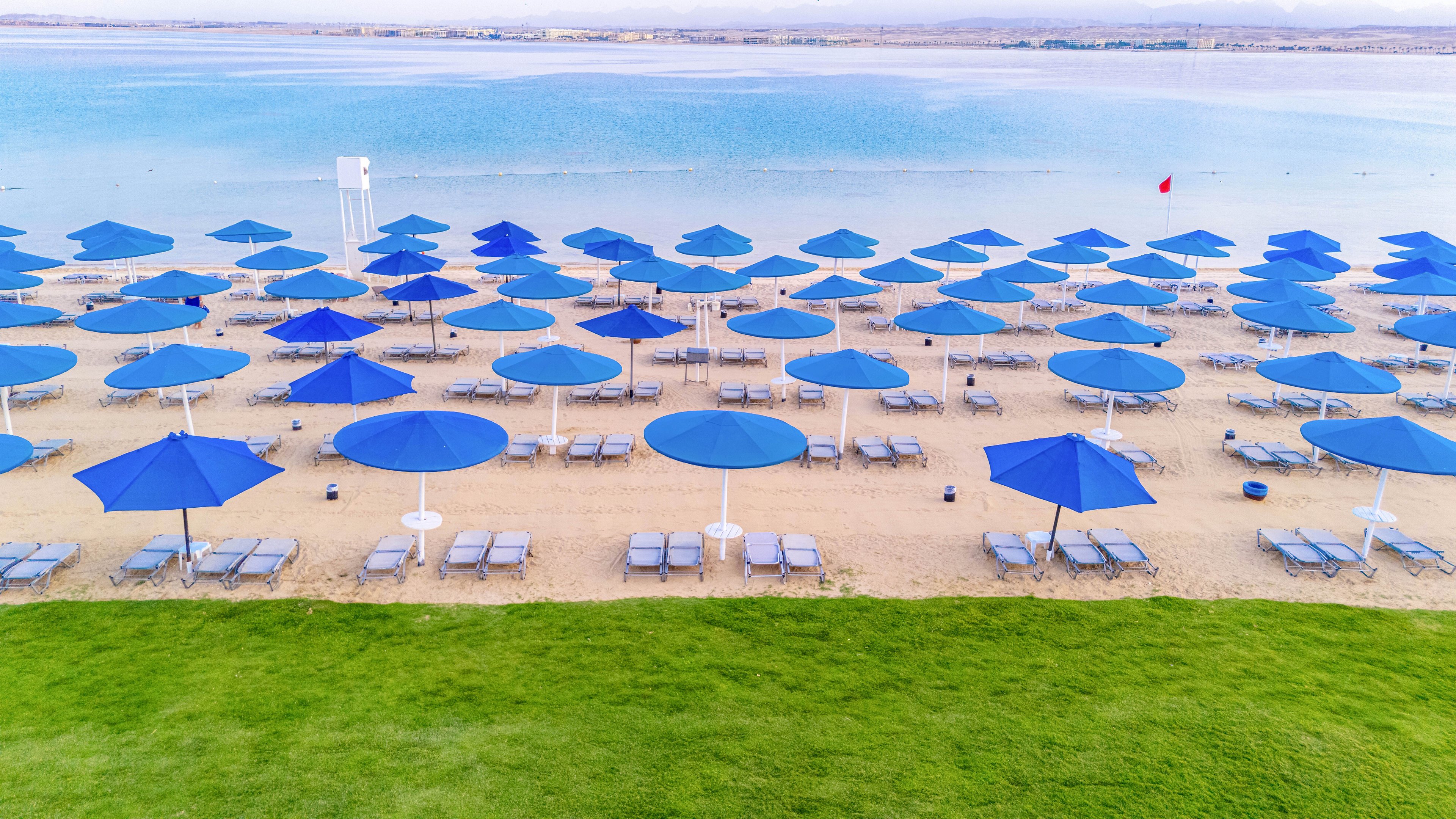 Rows of blue beach umbrellas and lounge chairs are neatly arranged on a sandy shoreline, facing a calm body of water. The scene is viewed from above, with a vibrant green grass foreground and tranquil blue water in the background. The setting appears organized and serene, evoking a relaxed vacation mood. No people are visible, emphasizing the peaceful and inviting atmosphere.