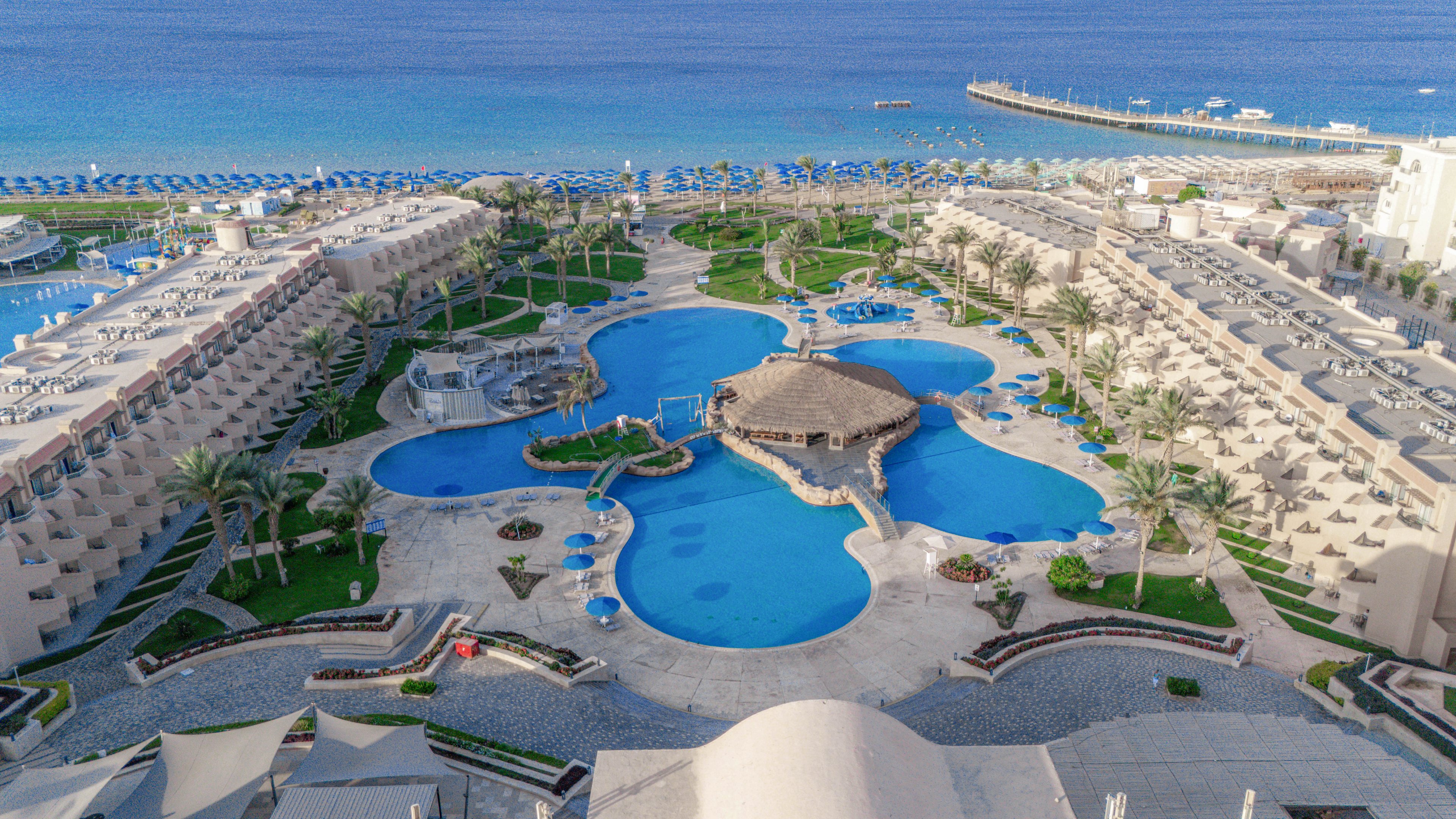 A large, uniquely shaped swimming pool is the central feature of this beachfront resort, surrounded by palm trees and sun loungers. The setting includes multiple buildings with balconies, a thatched-roof pool bar, and a clear view of the blue sea and pier in the background. The overall mood is tranquil and inviting, with a bright, sunny palette and well-maintained landscaping. No visible text or numbers are present in the image.