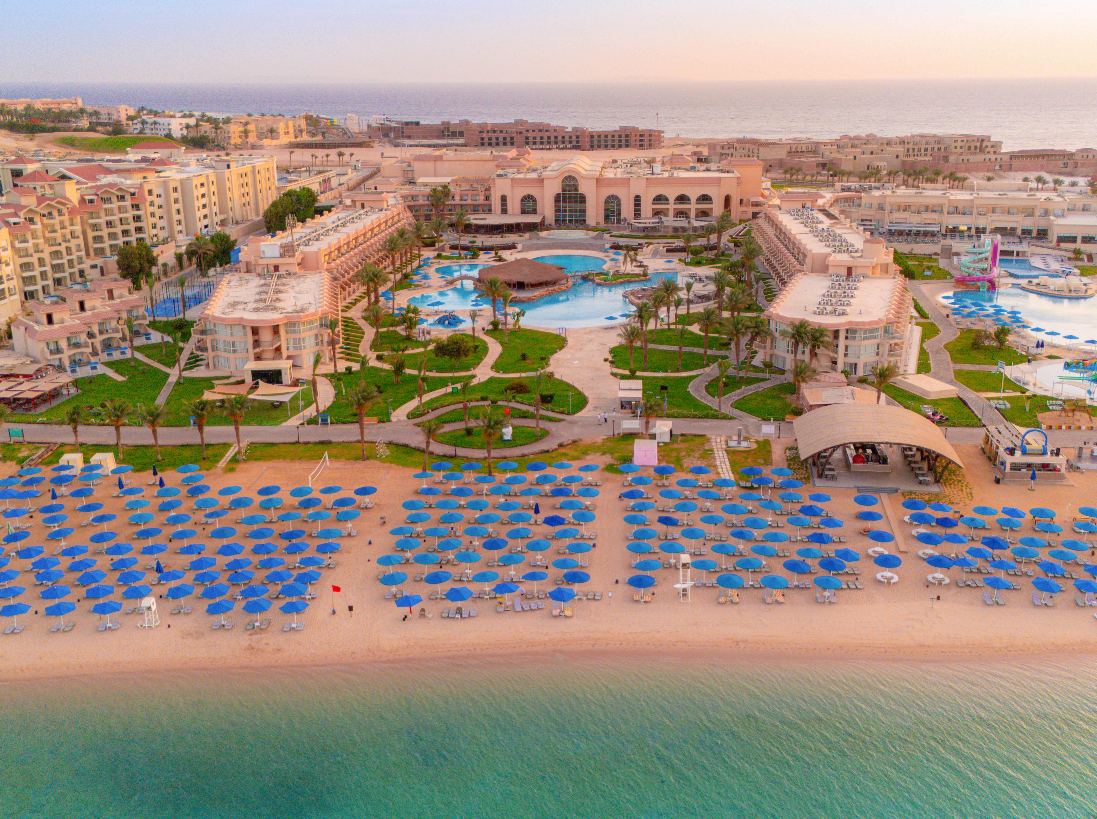 This image captures a sprawling luxury resort complex situated directly on a sandy beachfront. Rows of blue umbrellas line the shore, while multiple pools and landscaped gardens are visible within the resort grounds. The architecture features grand facades and palm trees, with the tranquil sea in the foreground and a pastel sky at sunrise. No visible text or numbers are present in the image.