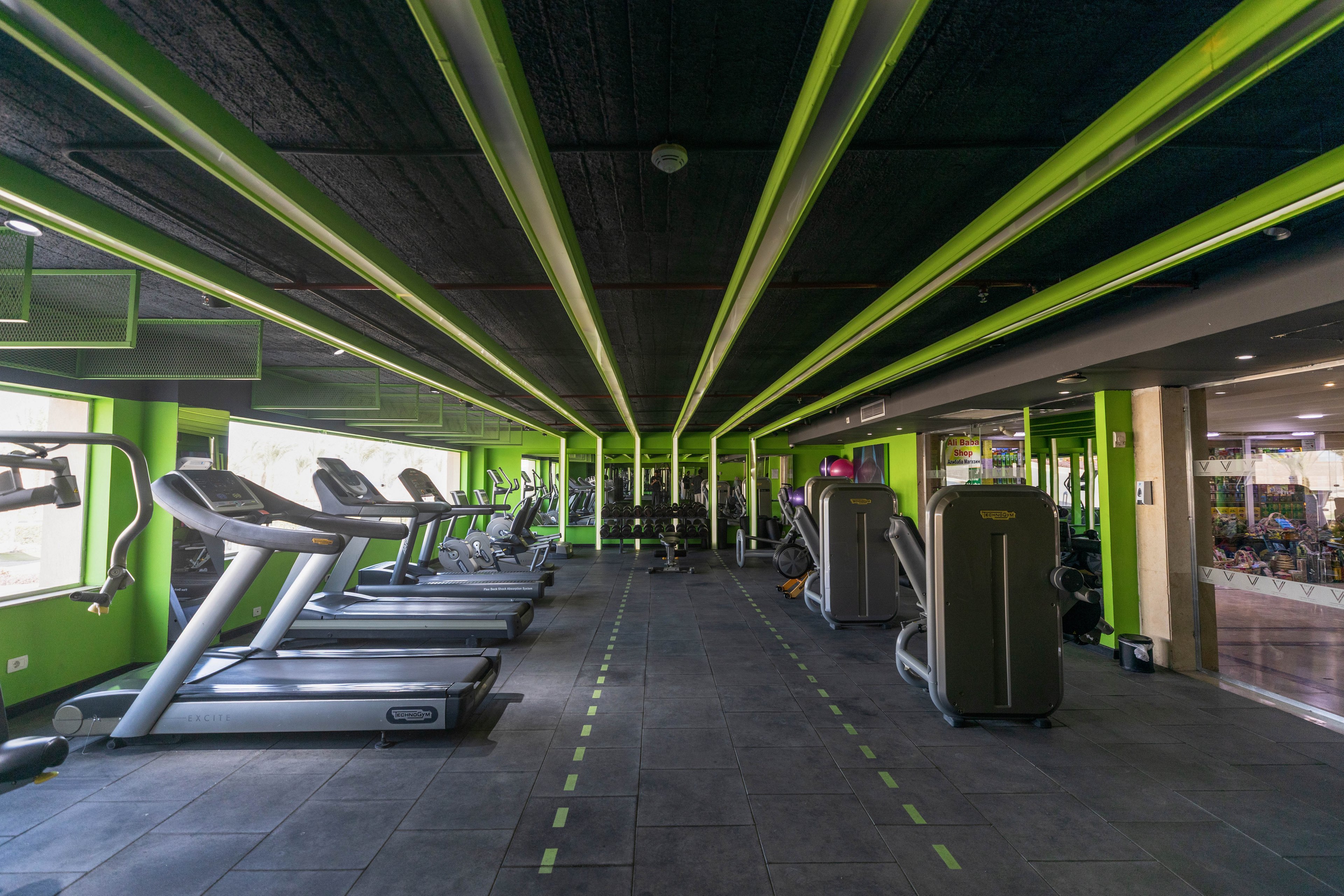 A spacious gym features multiple treadmills and exercise machines arranged in neat rows. The setting is brightly lit with green accents on the walls and ceiling, creating a vibrant and energetic atmosphere. The gym appears empty, emphasizing the clean and organized environment. No visible text or numbers are present on the equipment or walls.