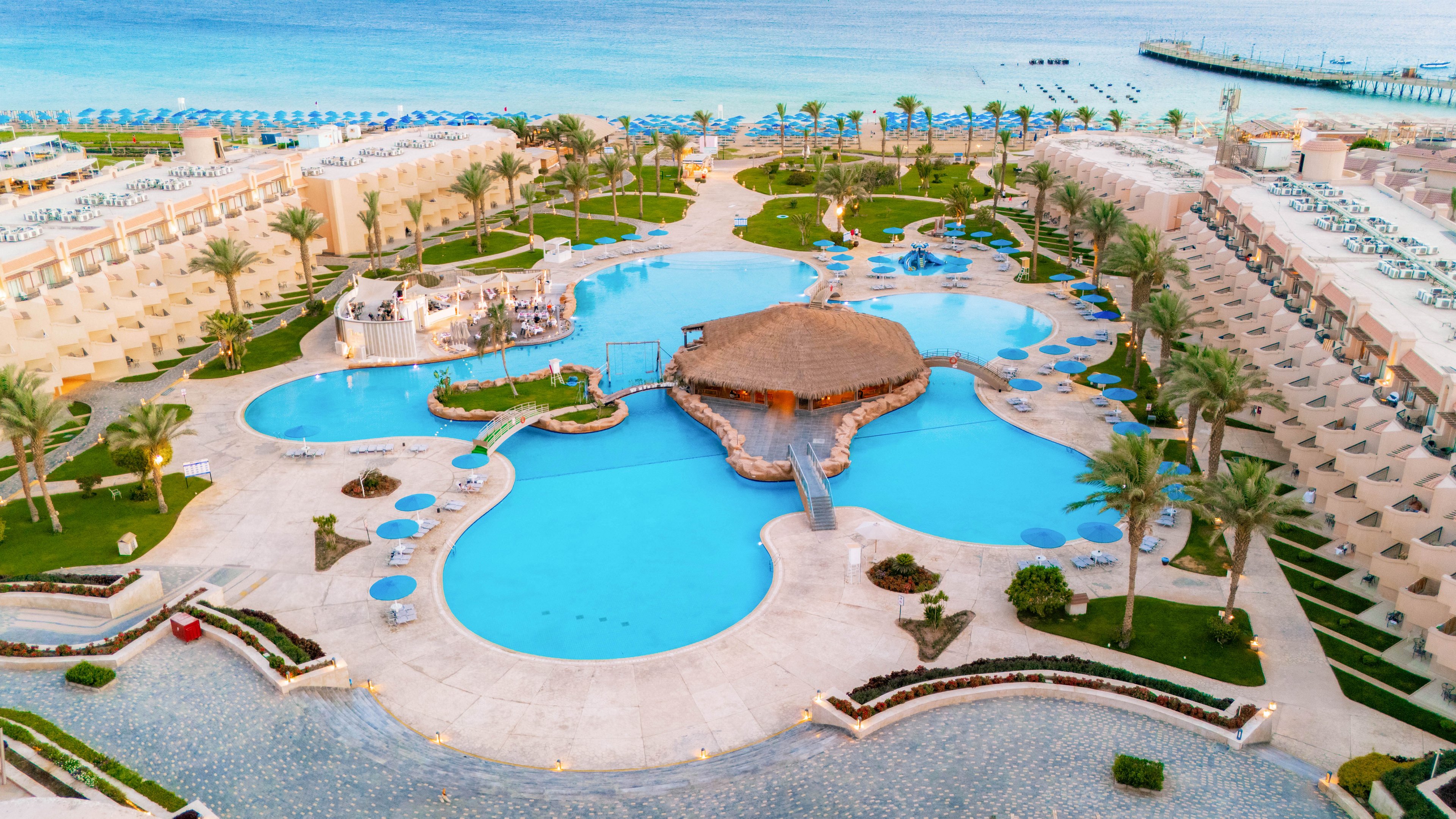A large, tropical resort complex features a central lagoon-style swimming pool surrounded by palm trees and sun loungers. The setting is beachfront, with the ocean and rows of umbrellas visible in the background. The architecture is symmetrical, with guest rooms lining both sides and a thatched-roof pool bar at the center. The mood is vibrant and relaxing, with a bright blue palette dominating the scene. No visible text or numbers are present in the image.