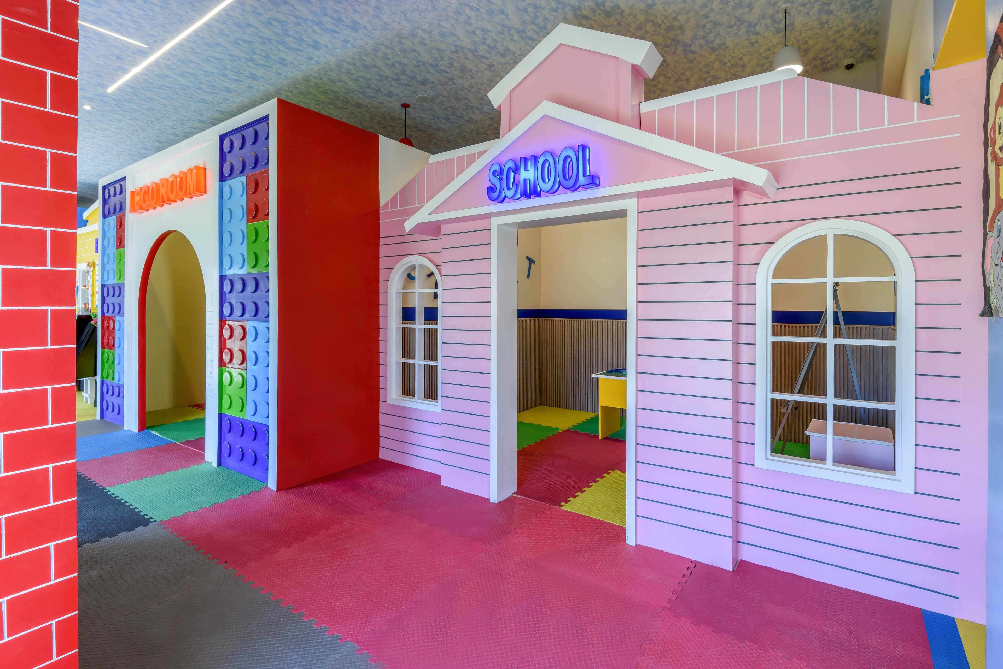 A vibrant indoor play space features a pink schoolhouse facade with a neon 'SCHOOL' sign above the entrance. The area includes large, colorful building block walls and soft foam flooring in various shades. The playful setting is designed for children, with bright lighting and imaginative structures encouraging creative play.